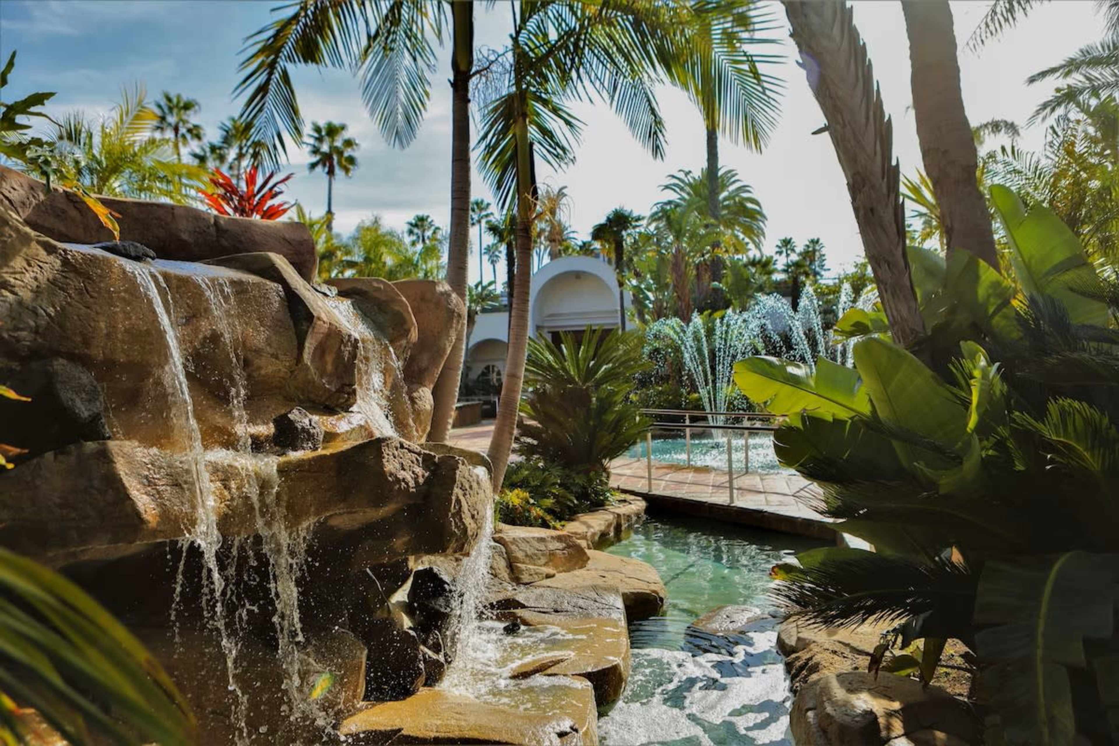 A waterfall cascades down rocky formations surrounded by palm trees and lush greenery, leading to a pool area in the background.