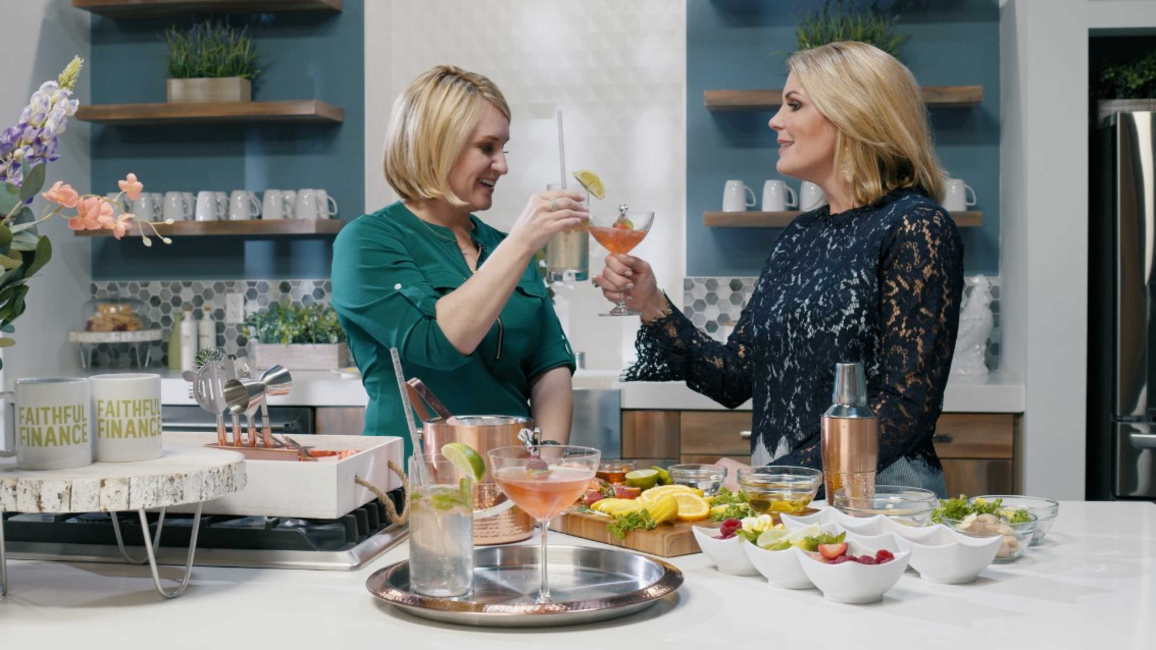 Two women are clinking glasses filled with cocktails in a kitchen, surrounded by various fruits and drinks on the counter.