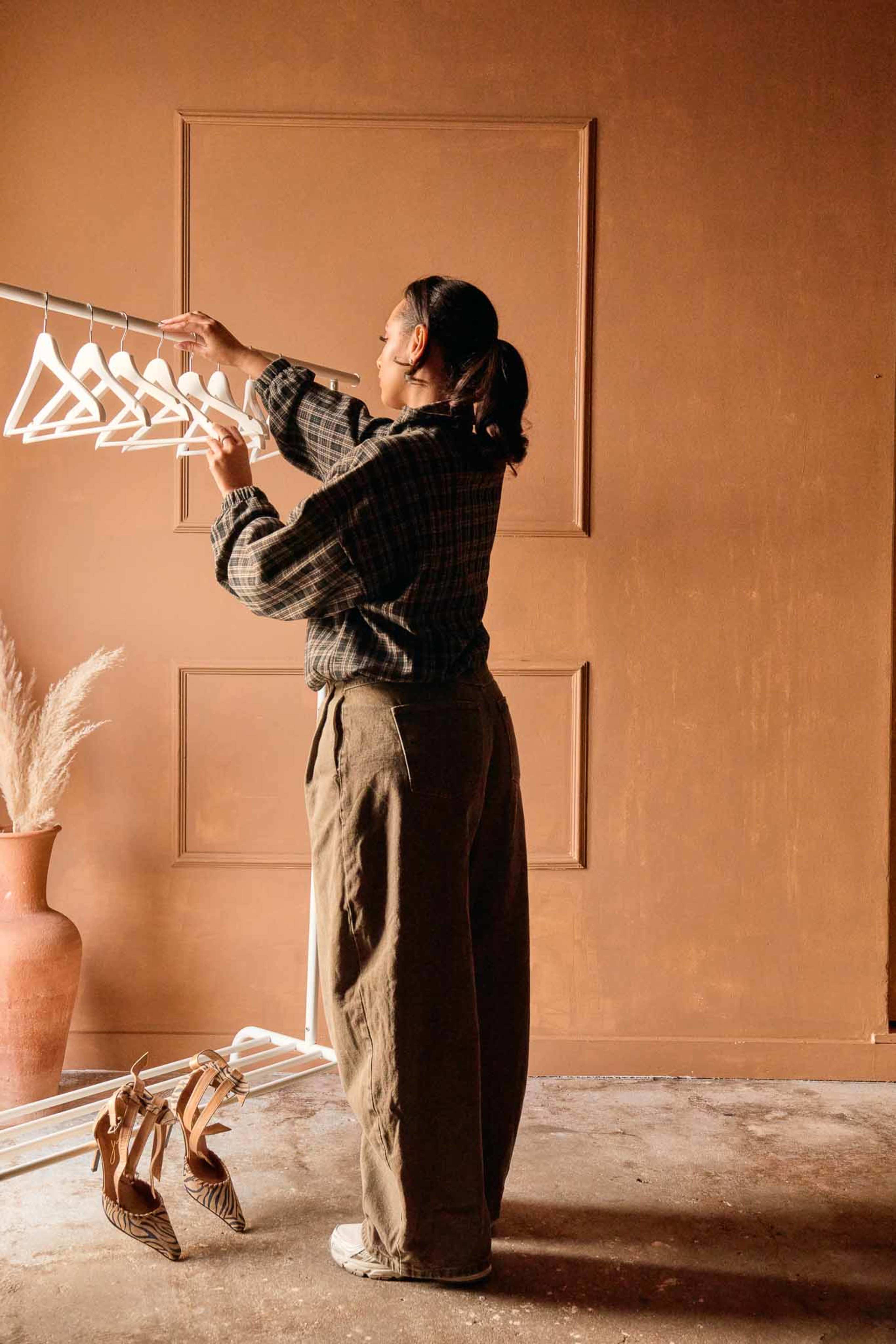 A person wearing a checkered shirt and loose pants is adjusting clothing hangers on a rack in a room with a warm-toned wall and a vase with dried plants.