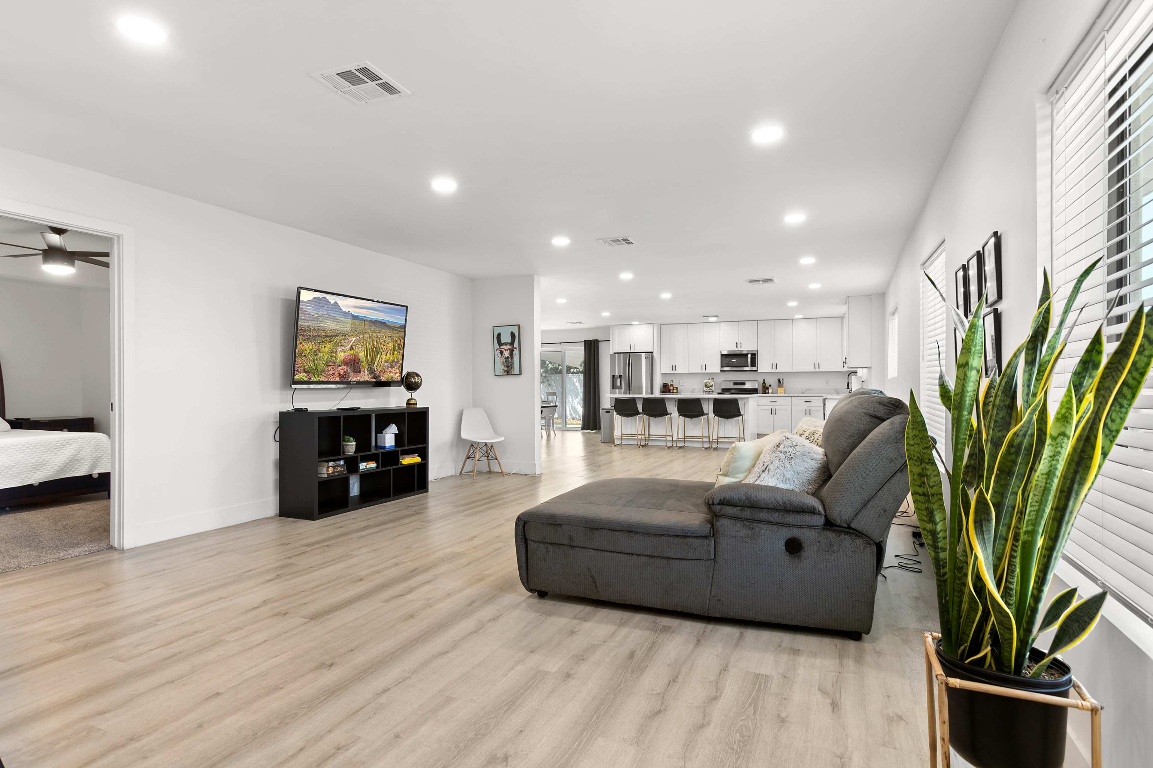 A modern living room features a grey sectional sofa facing a television mounted on the wall, with a view into an open kitchen area and plants in the foreground.