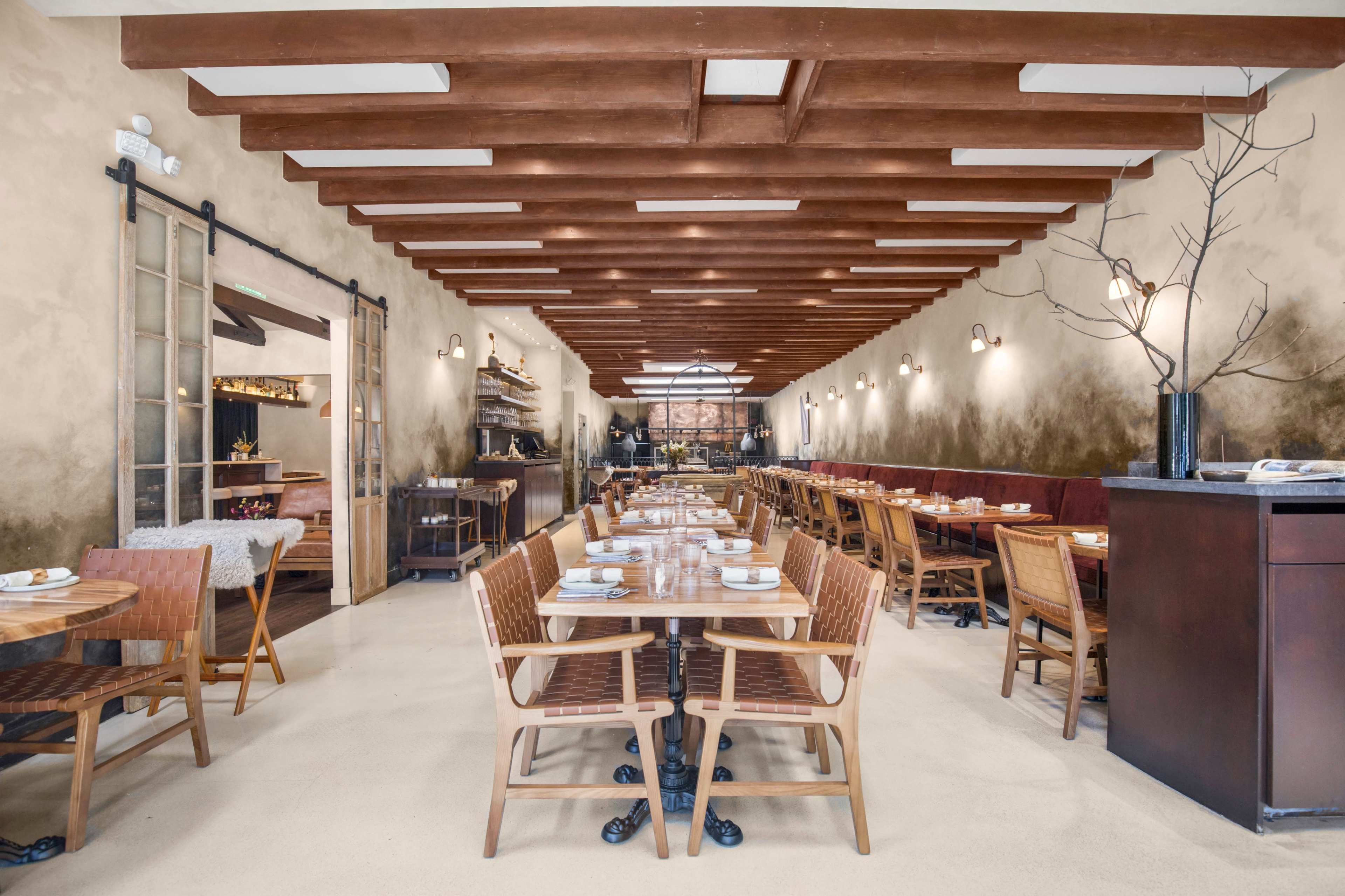 The image shows a modern restaurant interior with wooden beams, tables set for dining, and a spacious layout featuring a mixture of seating arrangements.