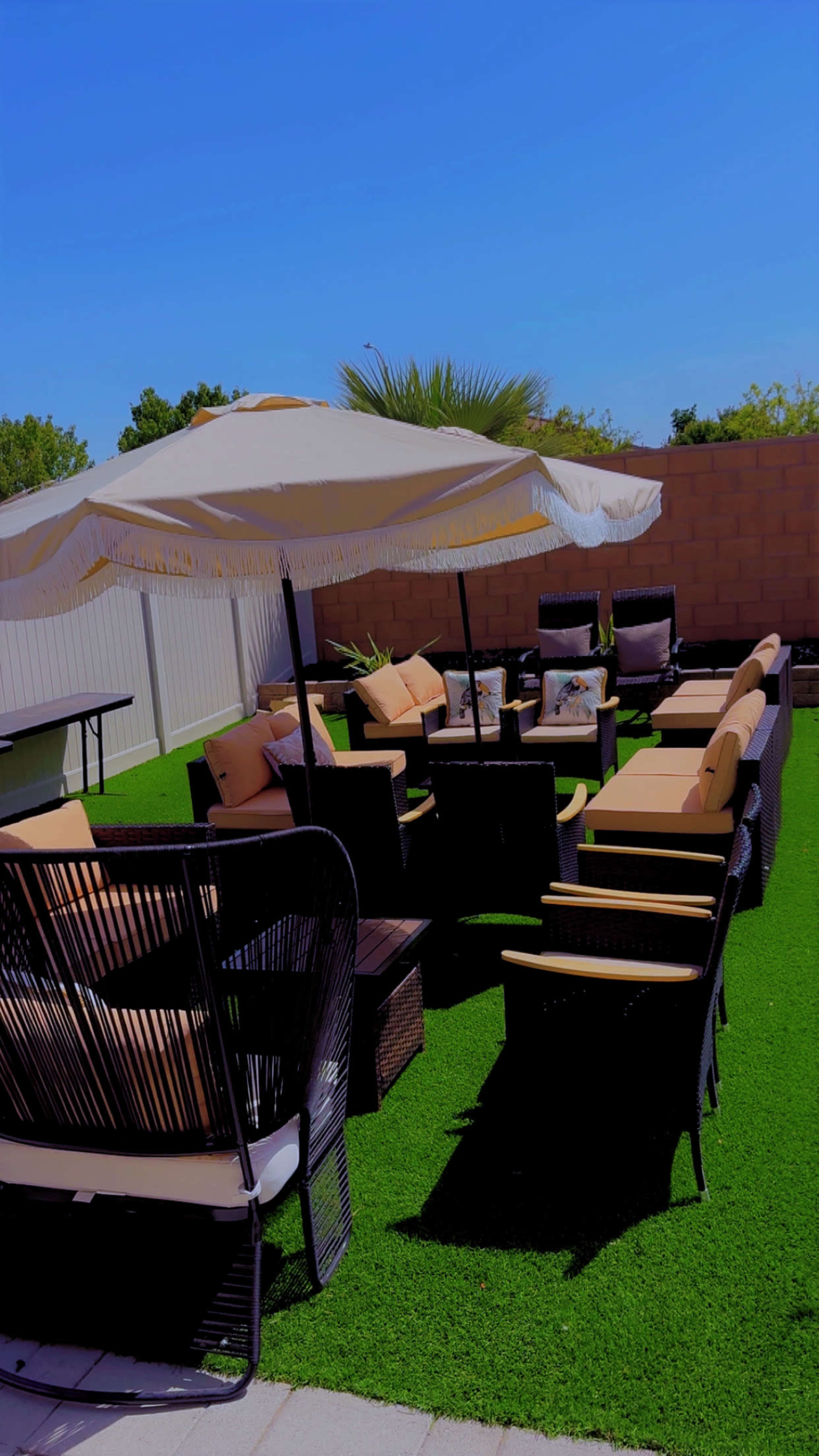 The image shows a patio area furnished with several black wicker chairs and couches, a large umbrella, and a table, all situated on green artificial grass.