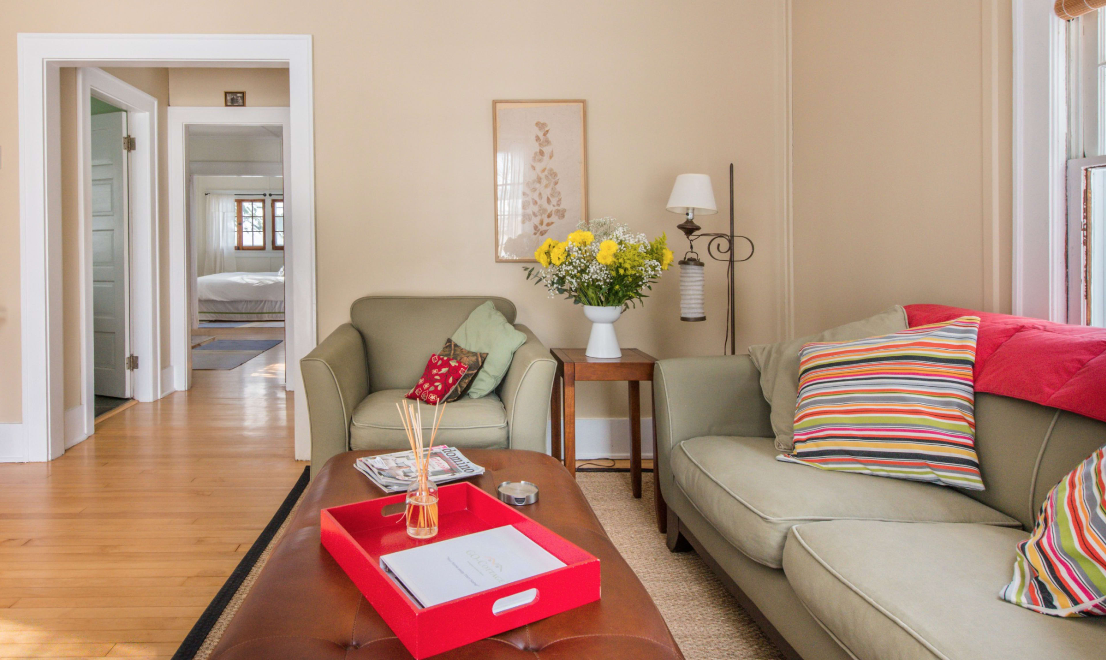 A cozy living room with a green sofa adorned with striped pillows, a wooden coffee table, and a vase of flowers on a side table, leading to a visible bedroom in the background.