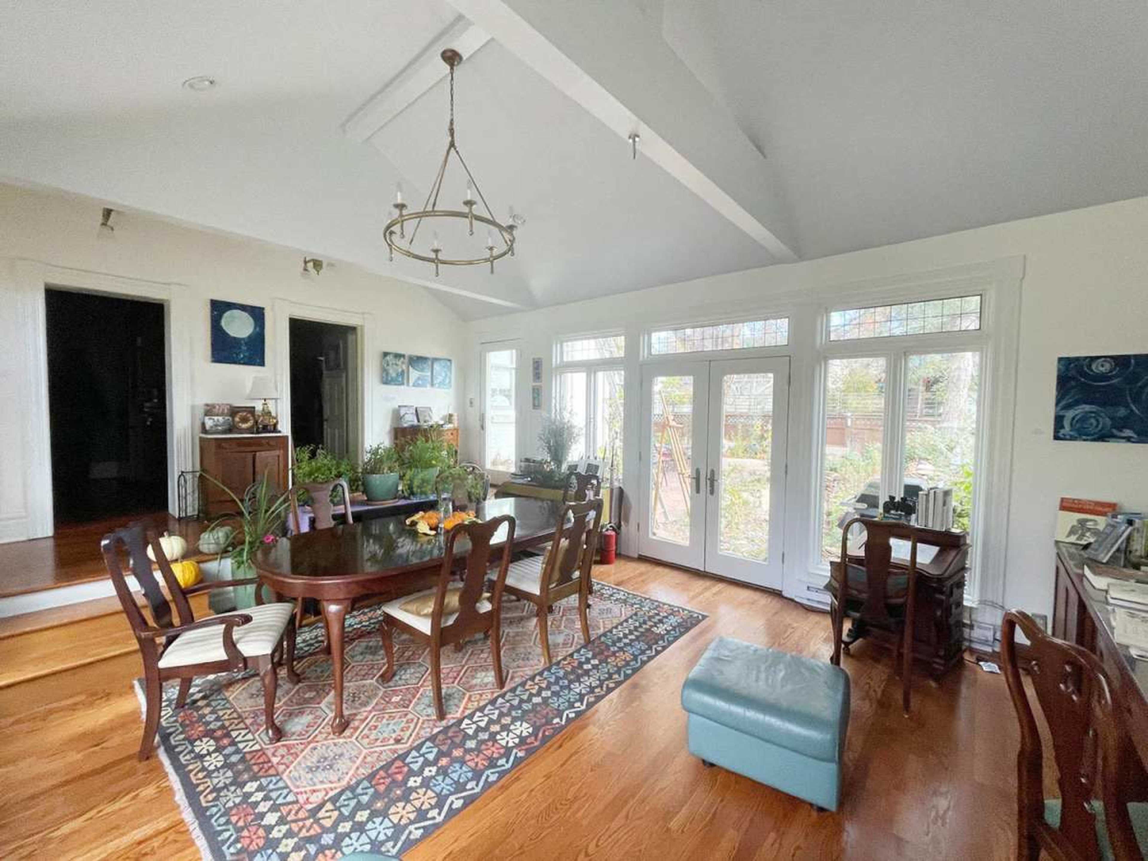 The image shows a spacious dining room with a large table surrounded by chairs, hardwood floors, and windows that provide natural light and outdoor views.