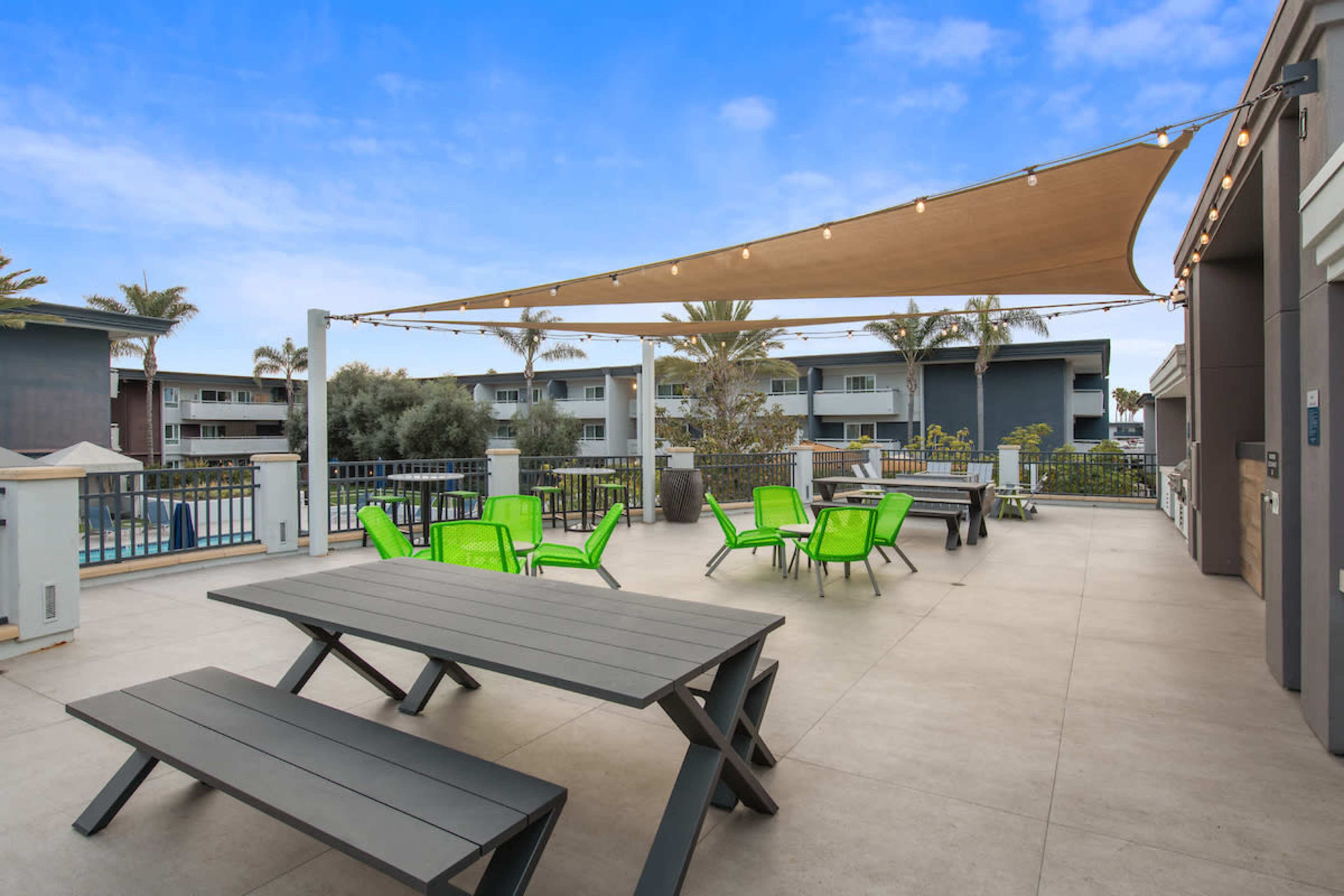 A patio area featuring a large picnic table, several bright green lounge chairs, and a sunshade, with buildings and palm trees in the background.