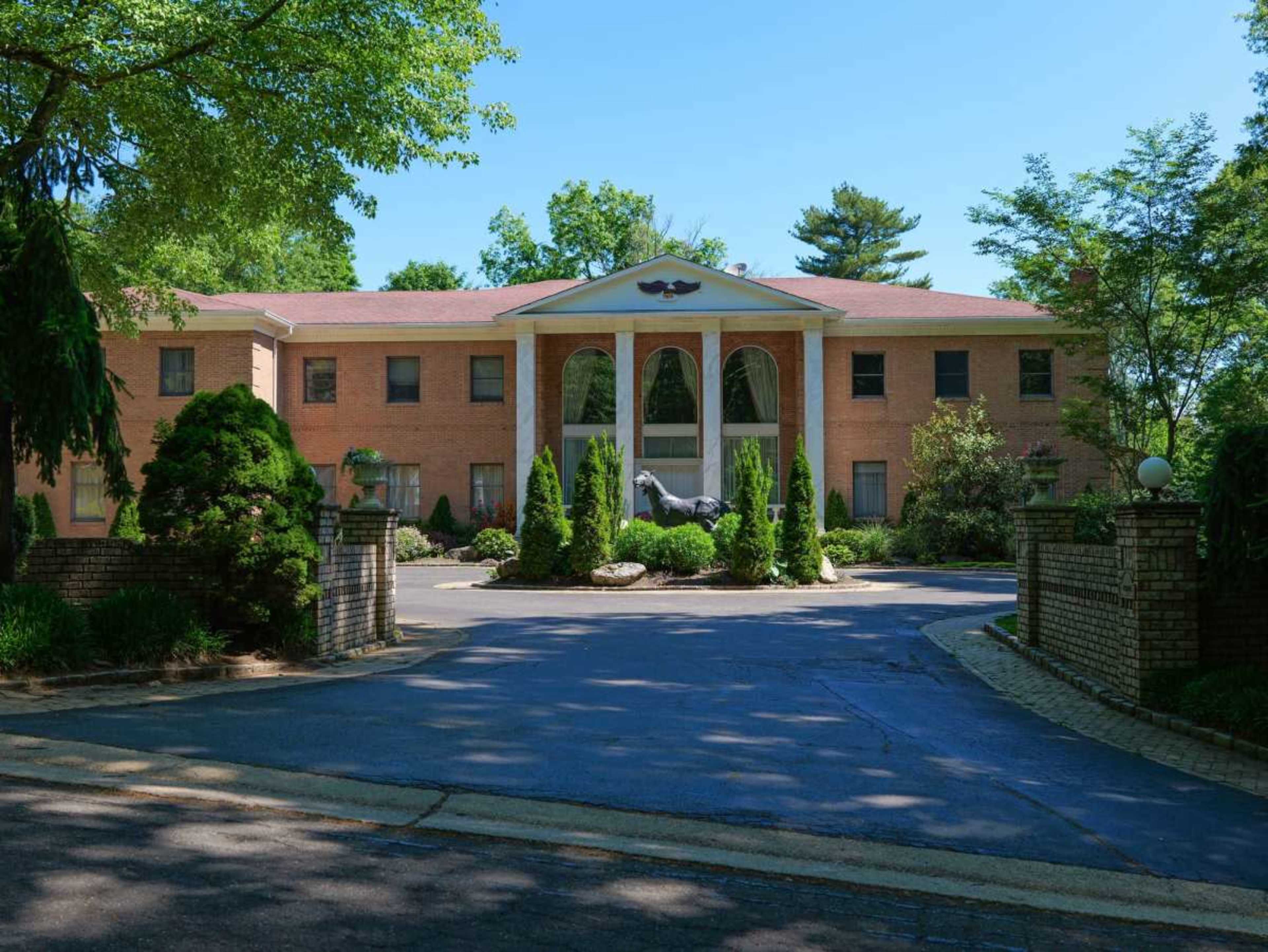 The image shows a large brick mansion with a central pediment supported by columns and flanked by manicured greenery in the front yard.