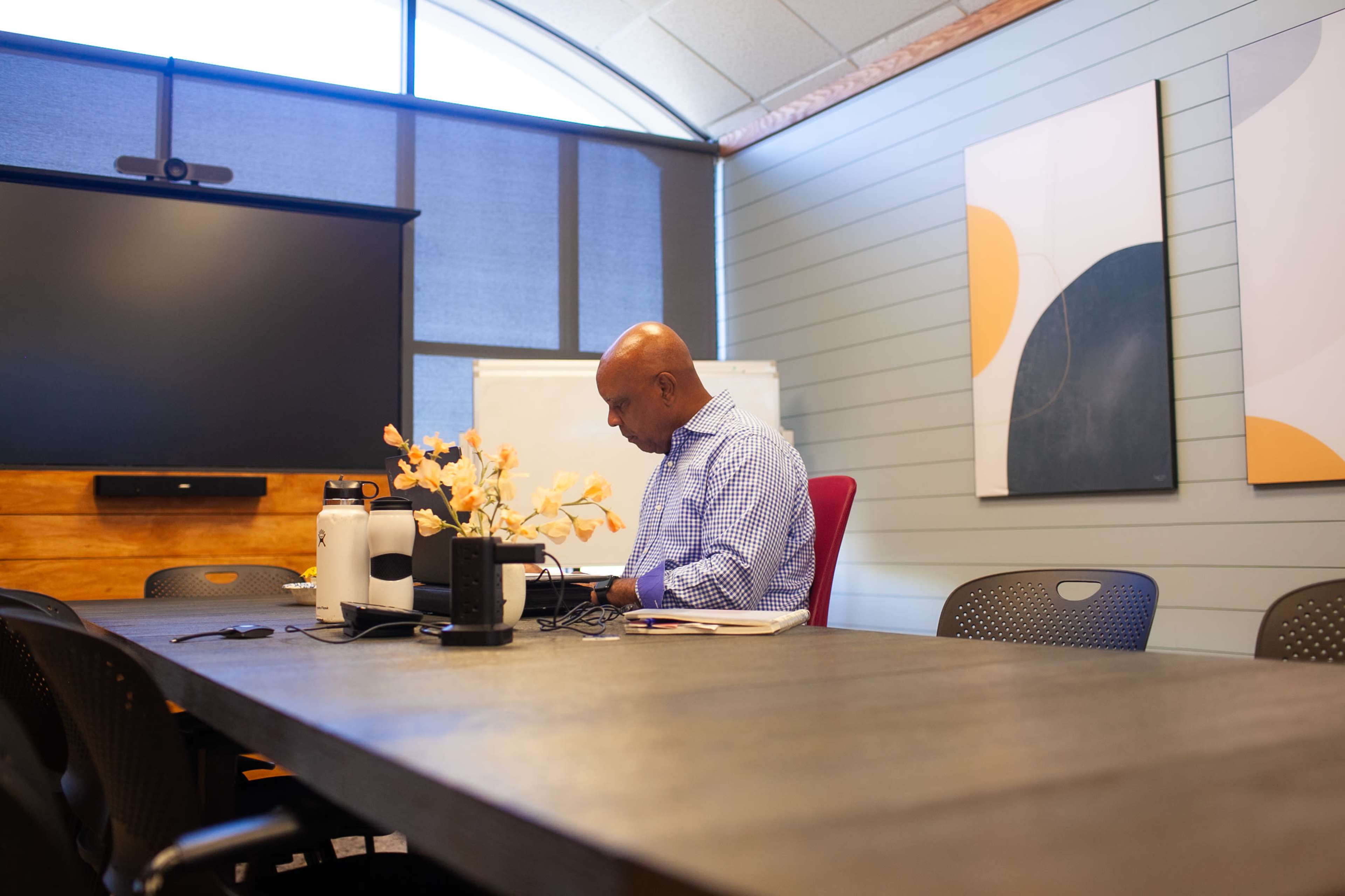 A man is seated at a conference table in a meeting room, working on a laptop with a floral arrangement in the center.