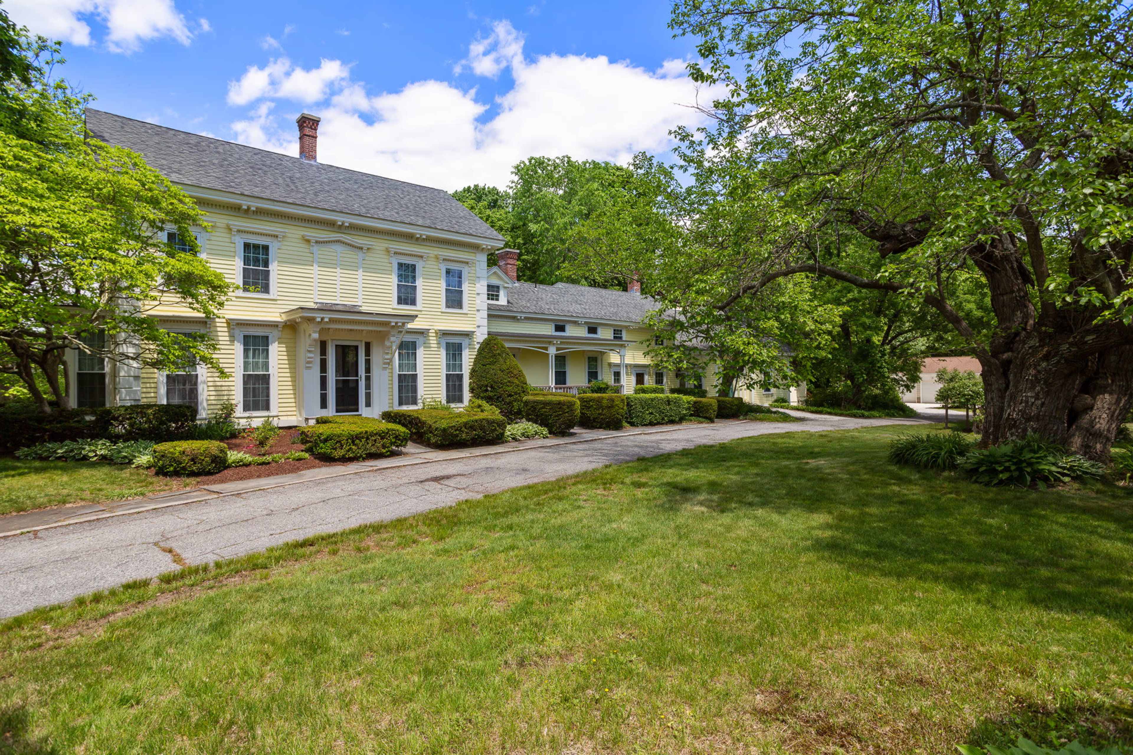 A yellow, two-story house with a porch and manicured landscaping is situated along a tree-lined driveway.