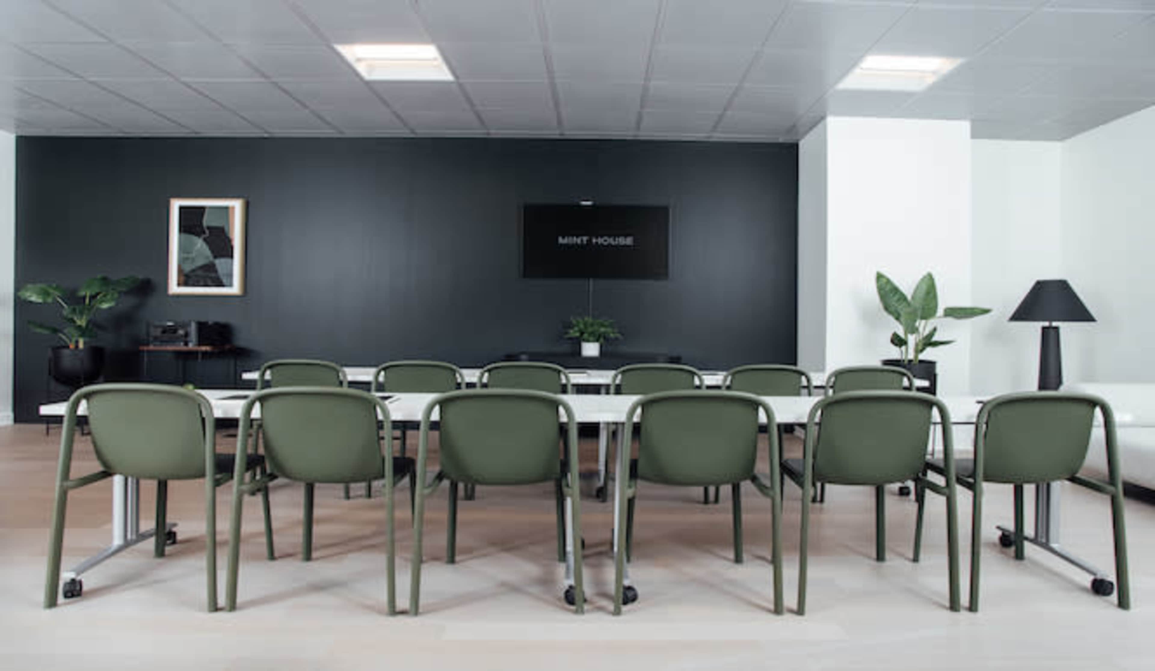 A modern conference room features a long table surrounded by green chairs, with a black wall that includes a sign labeled "MINT HOUSE."