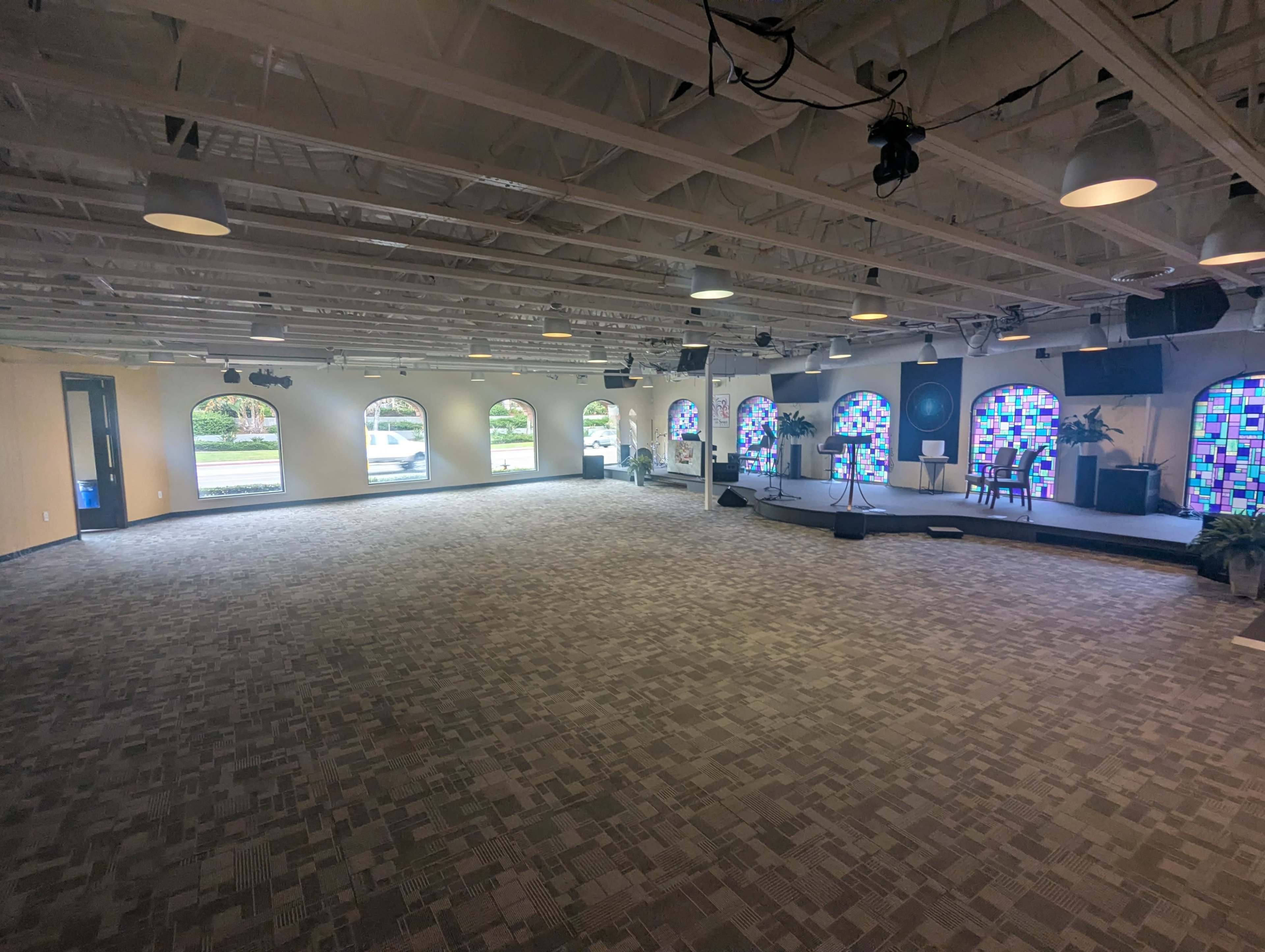 The image shows a spacious interior room with a carpeted floor, arched windows, and colorful stained glass panels, accompanied by lighting fixtures and minimal furniture.