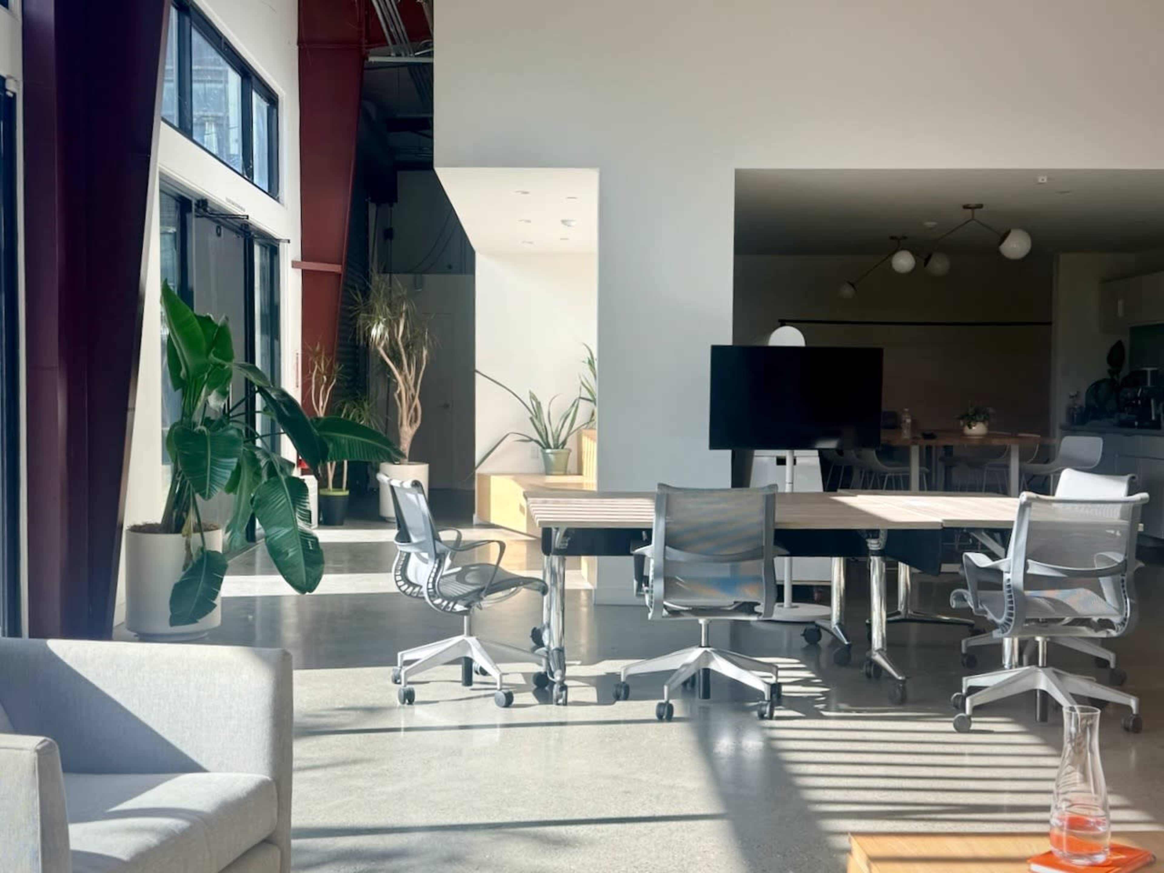 The image shows a modern conference room with a large table, office chairs, a digital screen, and large windows allowing natural light to enter.