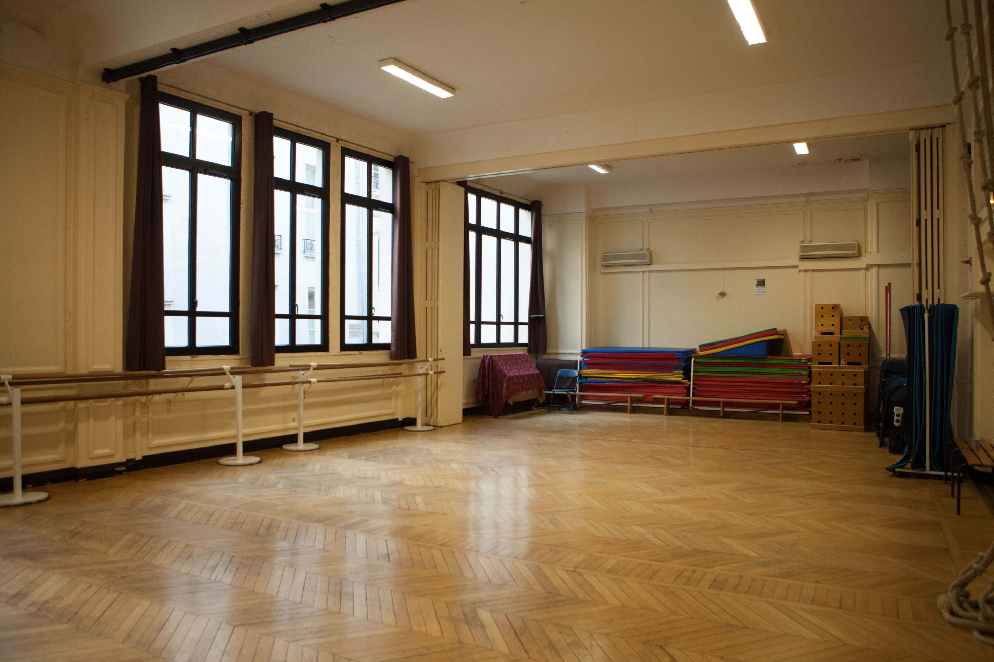 The image shows an empty room with wooden flooring, large windows, and various items including colorful benches and storage boxes along the walls.