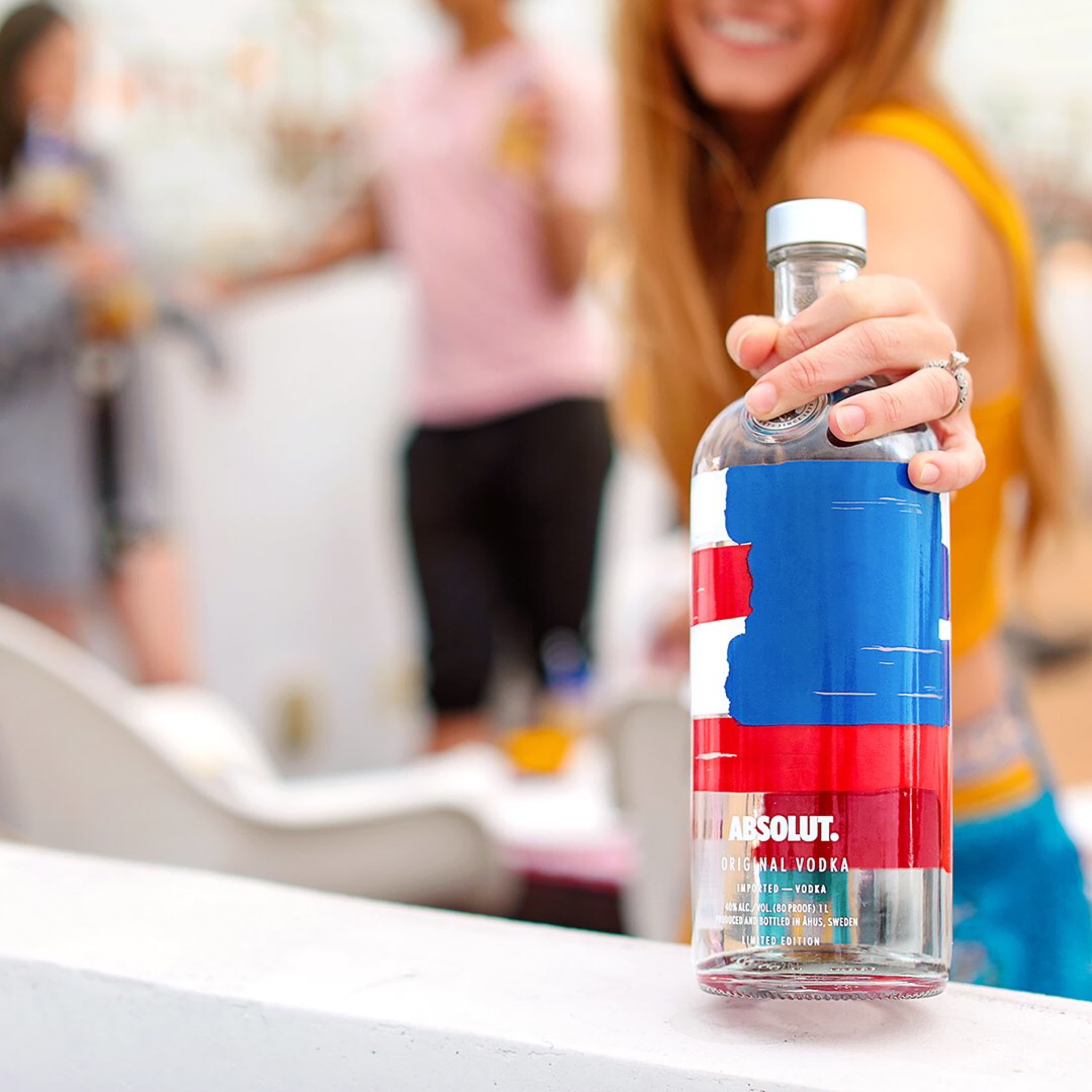 A woman holds a bottle of Absolut vodka with a blue and red design in a social setting.
