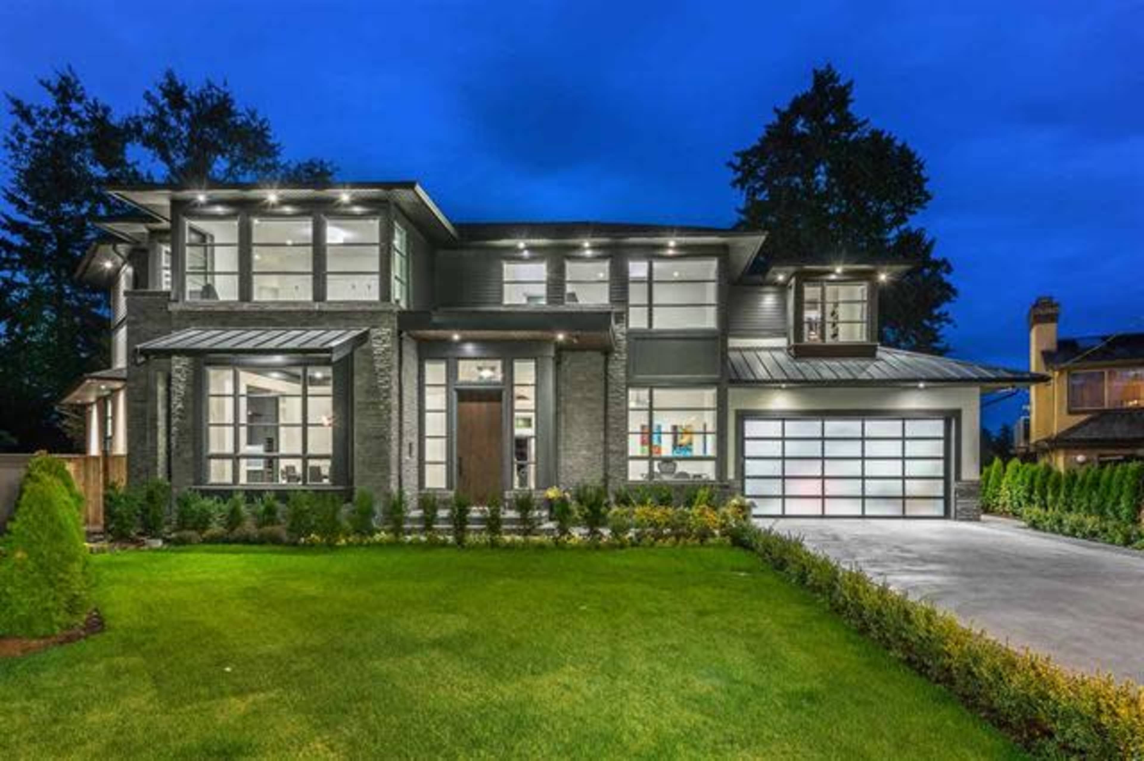 A modern two-story house with a dark exterior, large windows, and a well-manicured lawn at dusk.