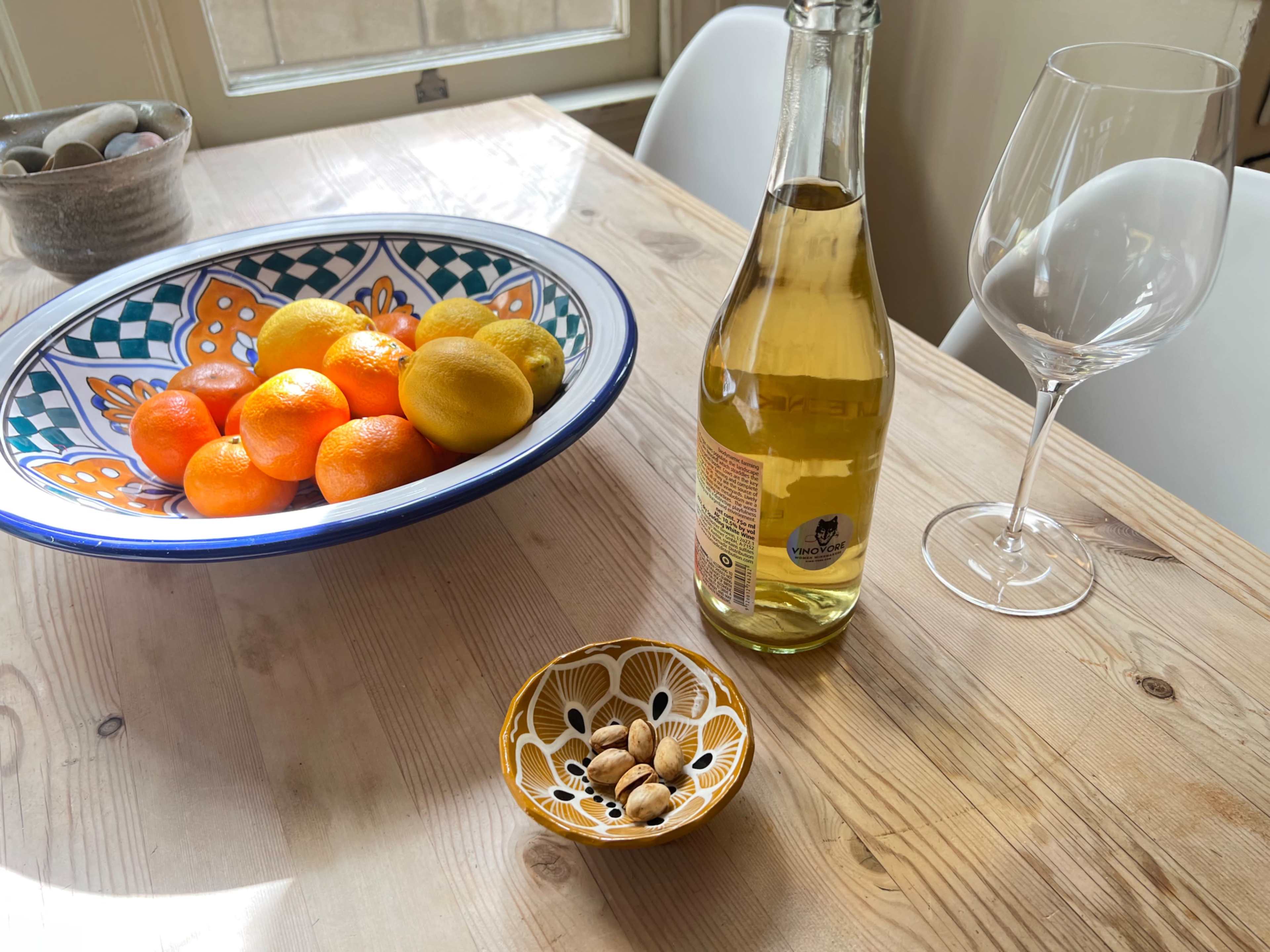 A bowl of citrus fruits sits on a wooden table beside a bottle of beverage and a dish of nuts.