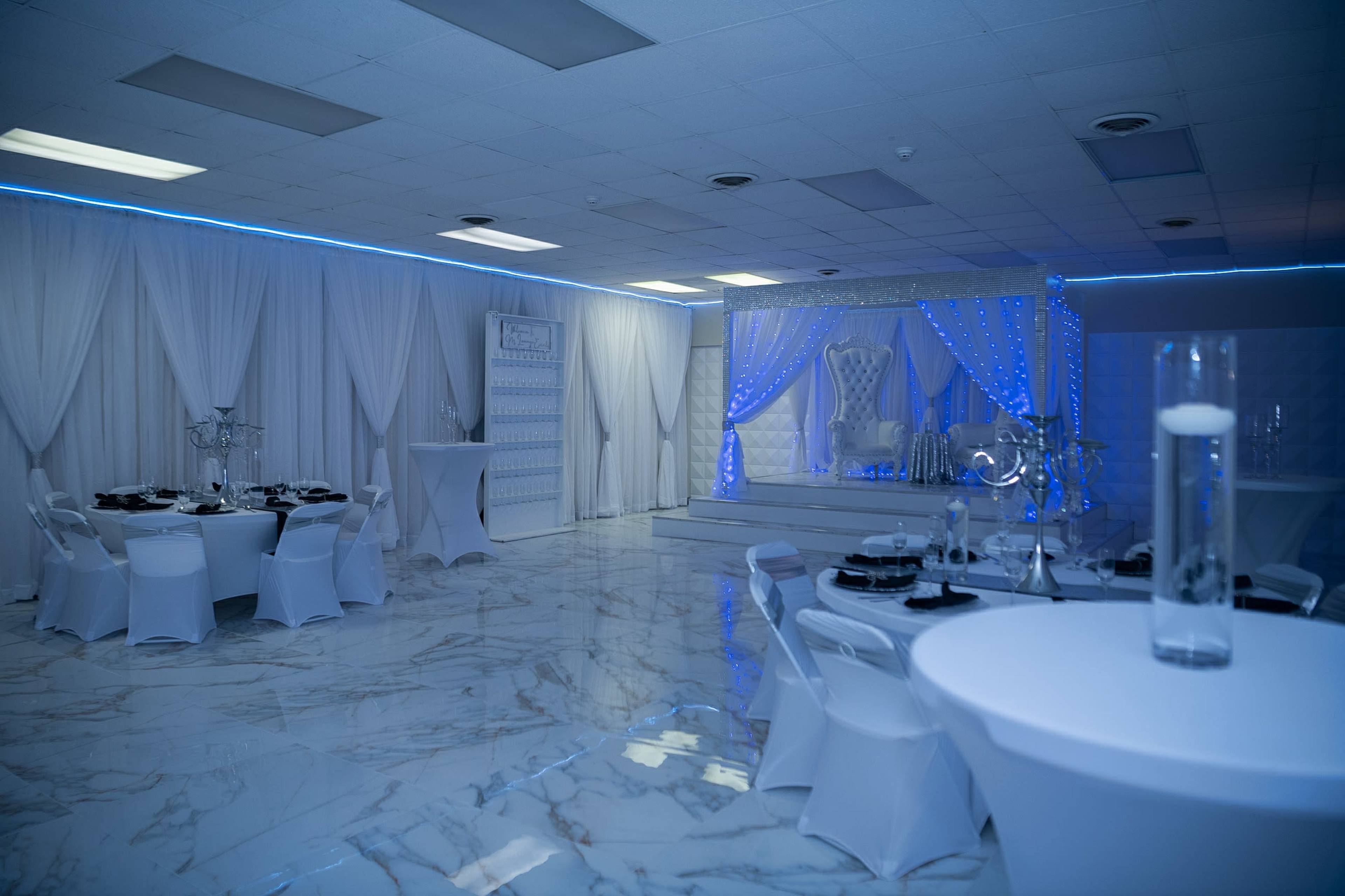 A spacious event hall features white table settings, elegant decor, and blue lighting, with a stage at the front adorned with drapes and a decorative throne.