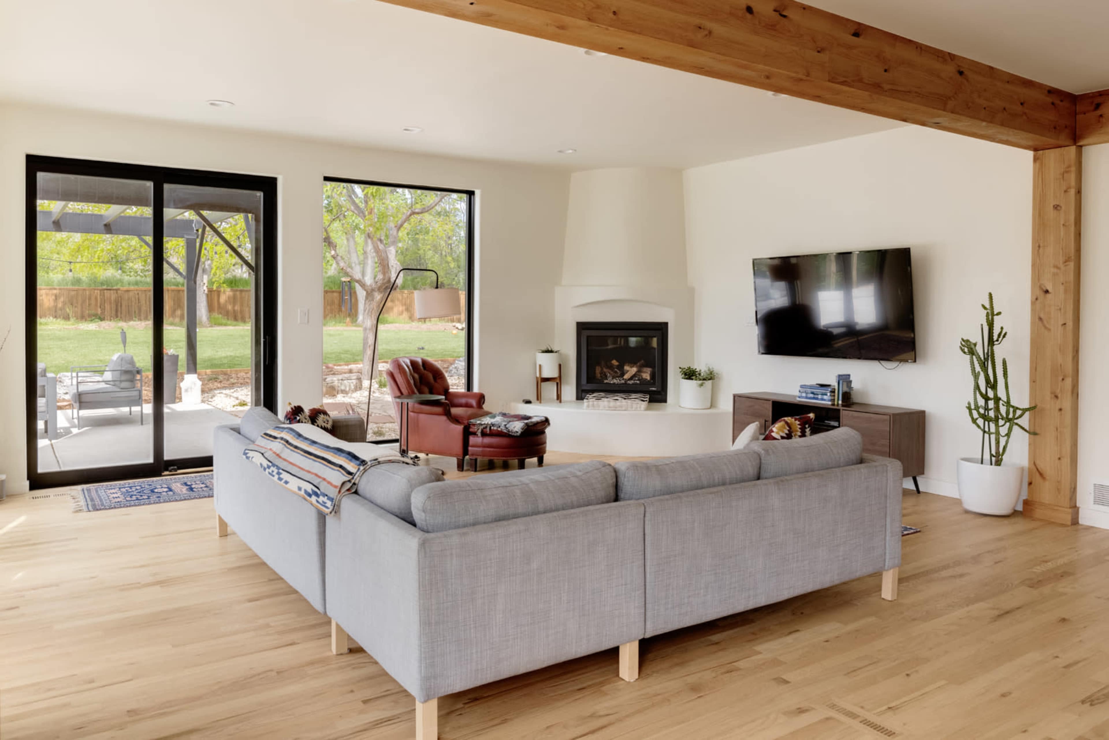 The image shows a modern living room with a gray sectional sofa, a fireplace, and large windows overlooking a backyard.