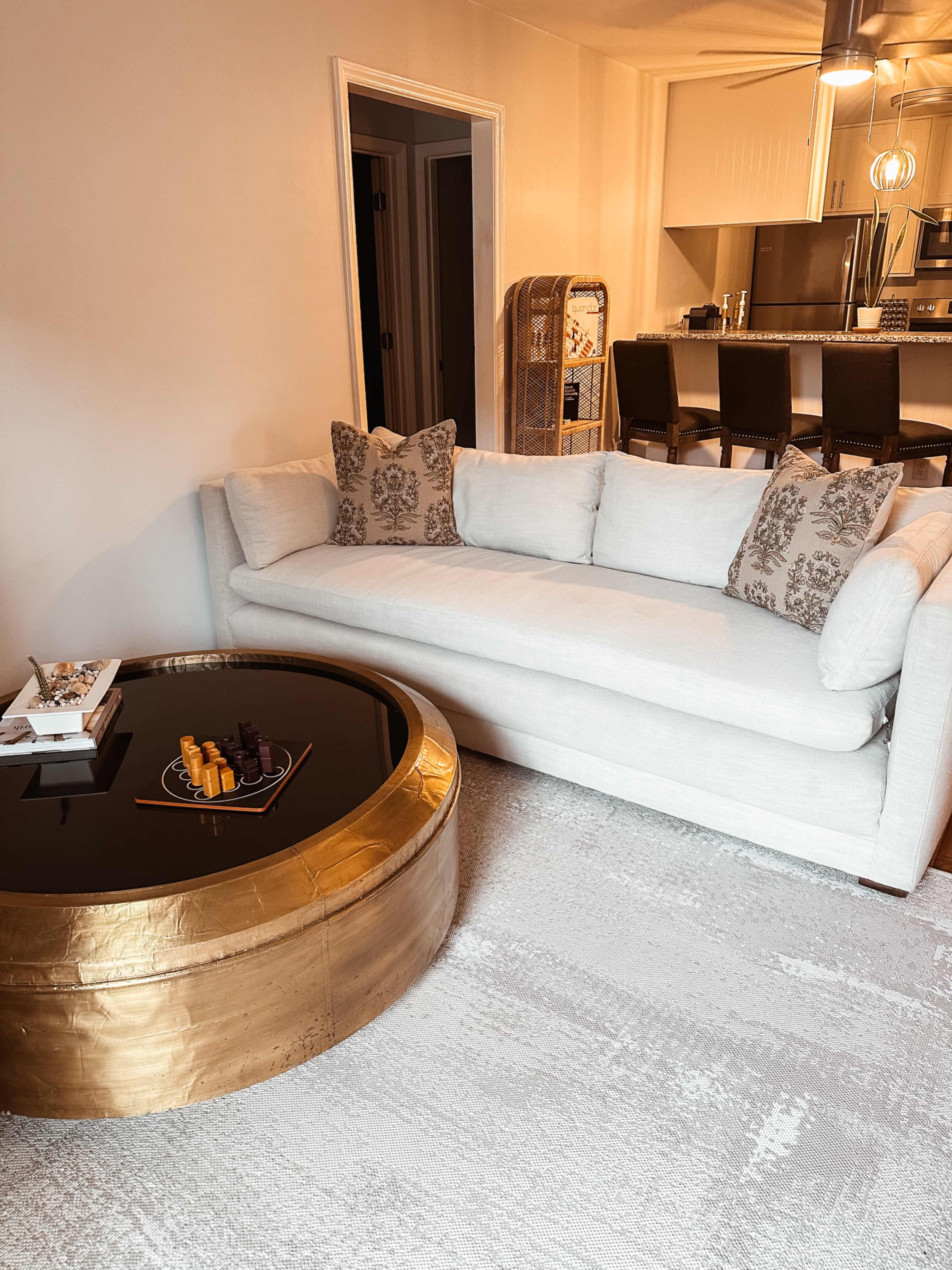 The image shows a light-colored sofa with decorative pillows, positioned next to a round coffee table with a black surface and a gold base, in a softly lit living area.