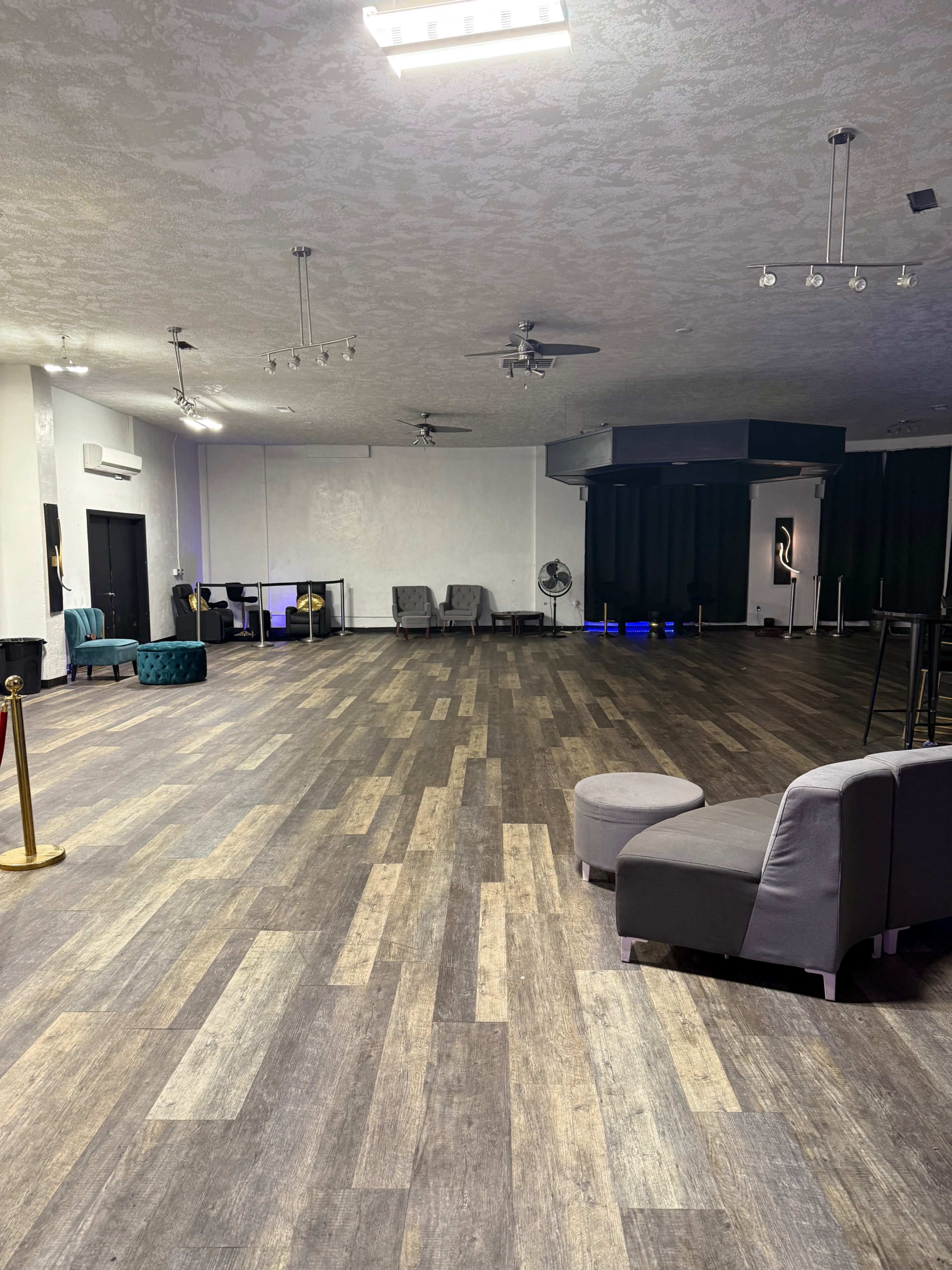 The image shows a spacious, empty room with a wooden floor, featuring a few chairs, a fan, and a stage area in the background.