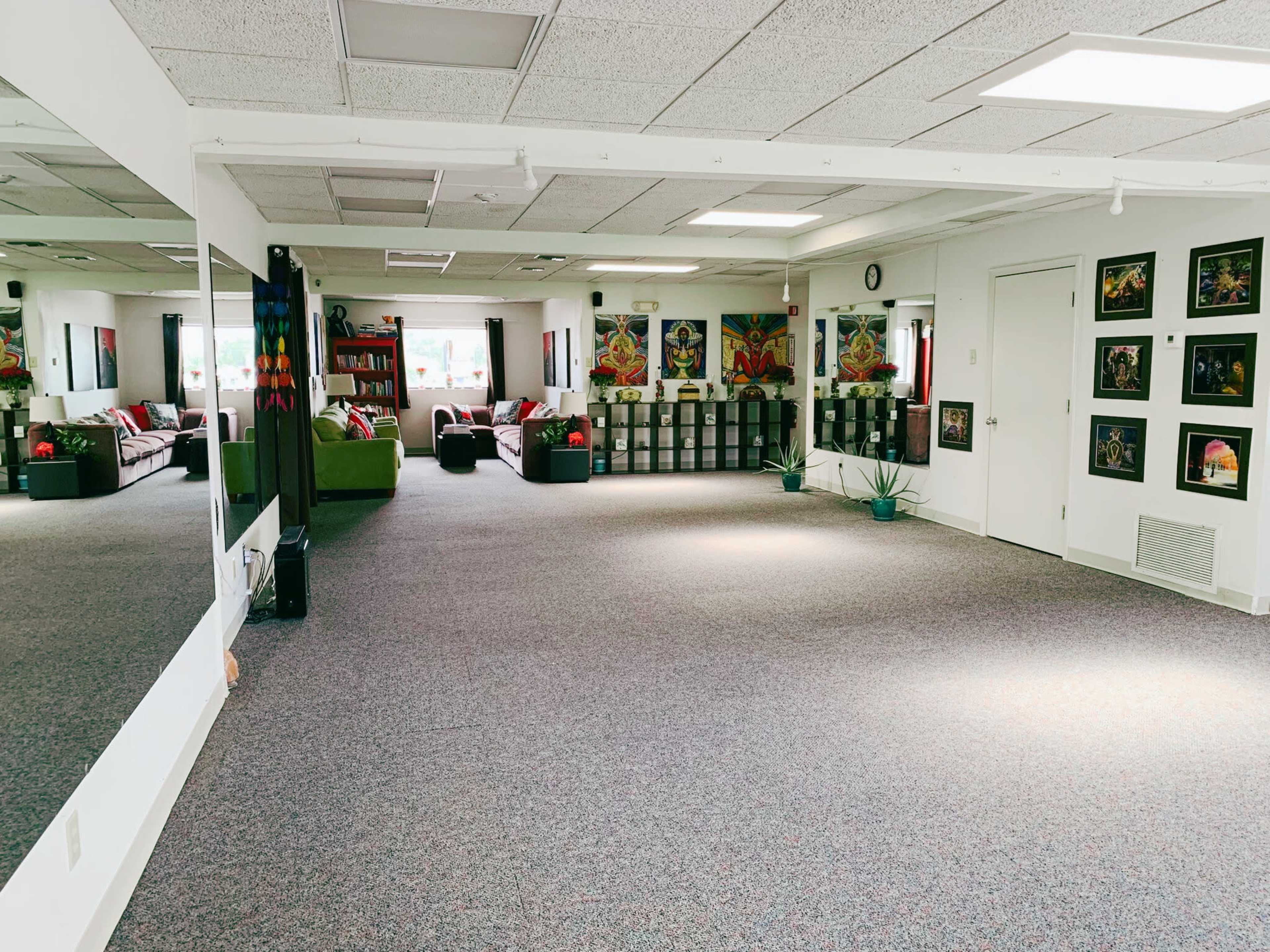A spacious, well-lit room with carpeted flooring, featuring a seating area with green couches, shelves filled with books, and framed artwork on the walls.