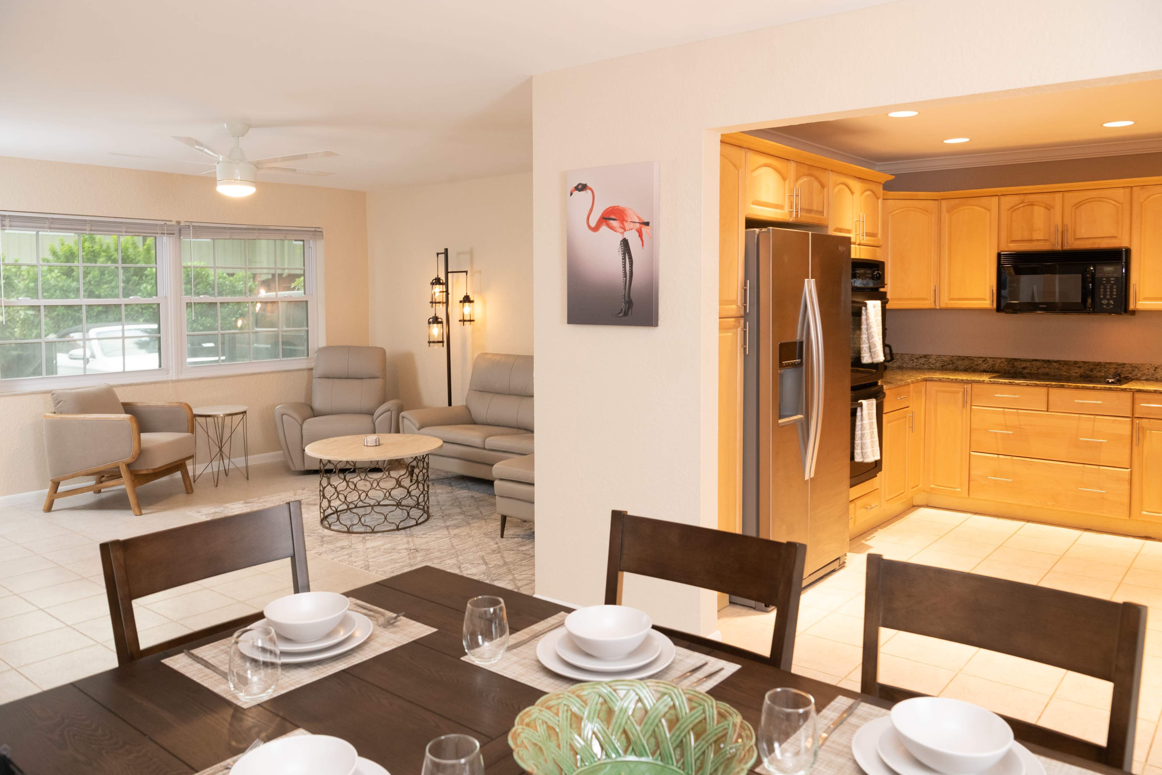 The image shows a modern kitchen and dining area with wooden cabinets, a table set for dinner, and a living space featuring comfortable seating and a wall-mounted artwork of a flamingo.