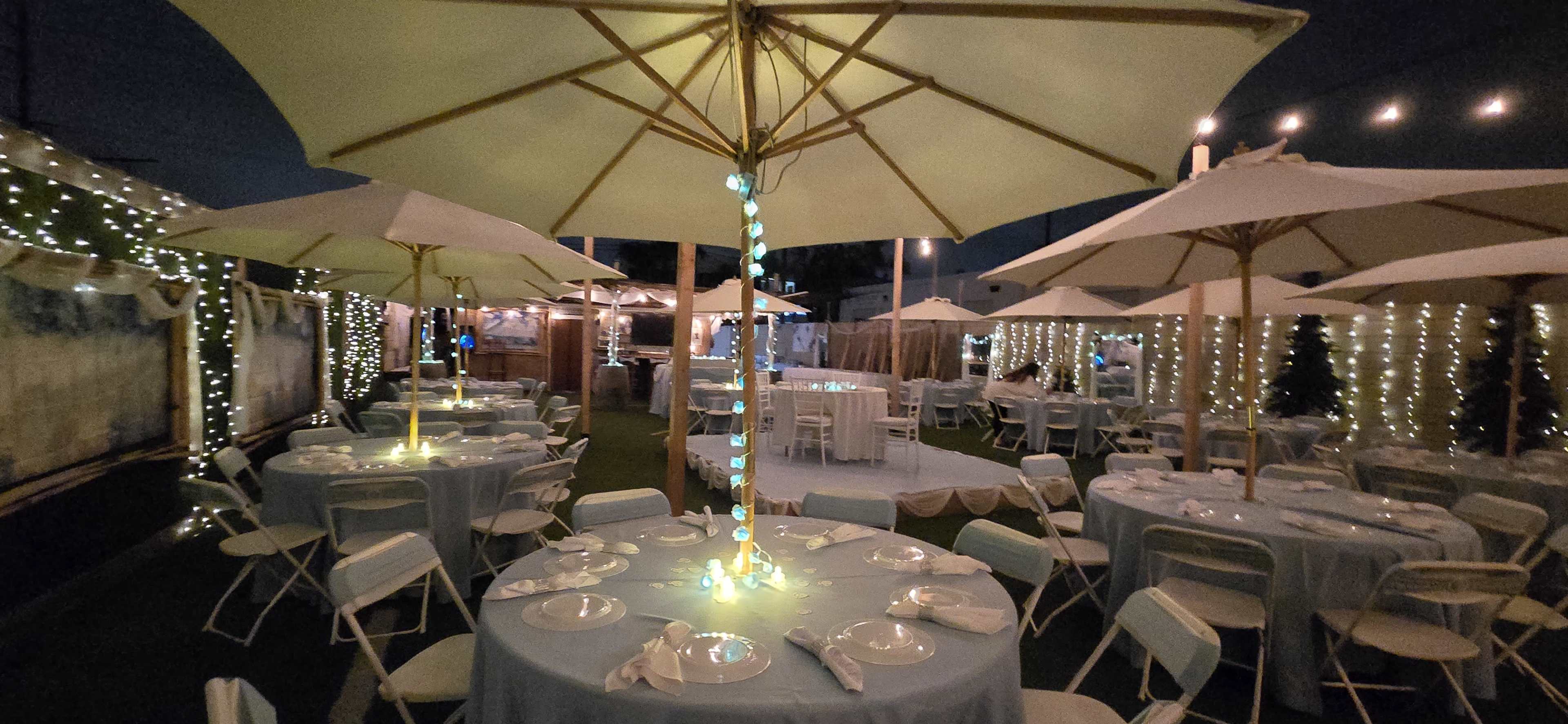 The scene features a decorated outdoor event space with tables and chairs set under large umbrellas, adorned with string lights and a soft glow.