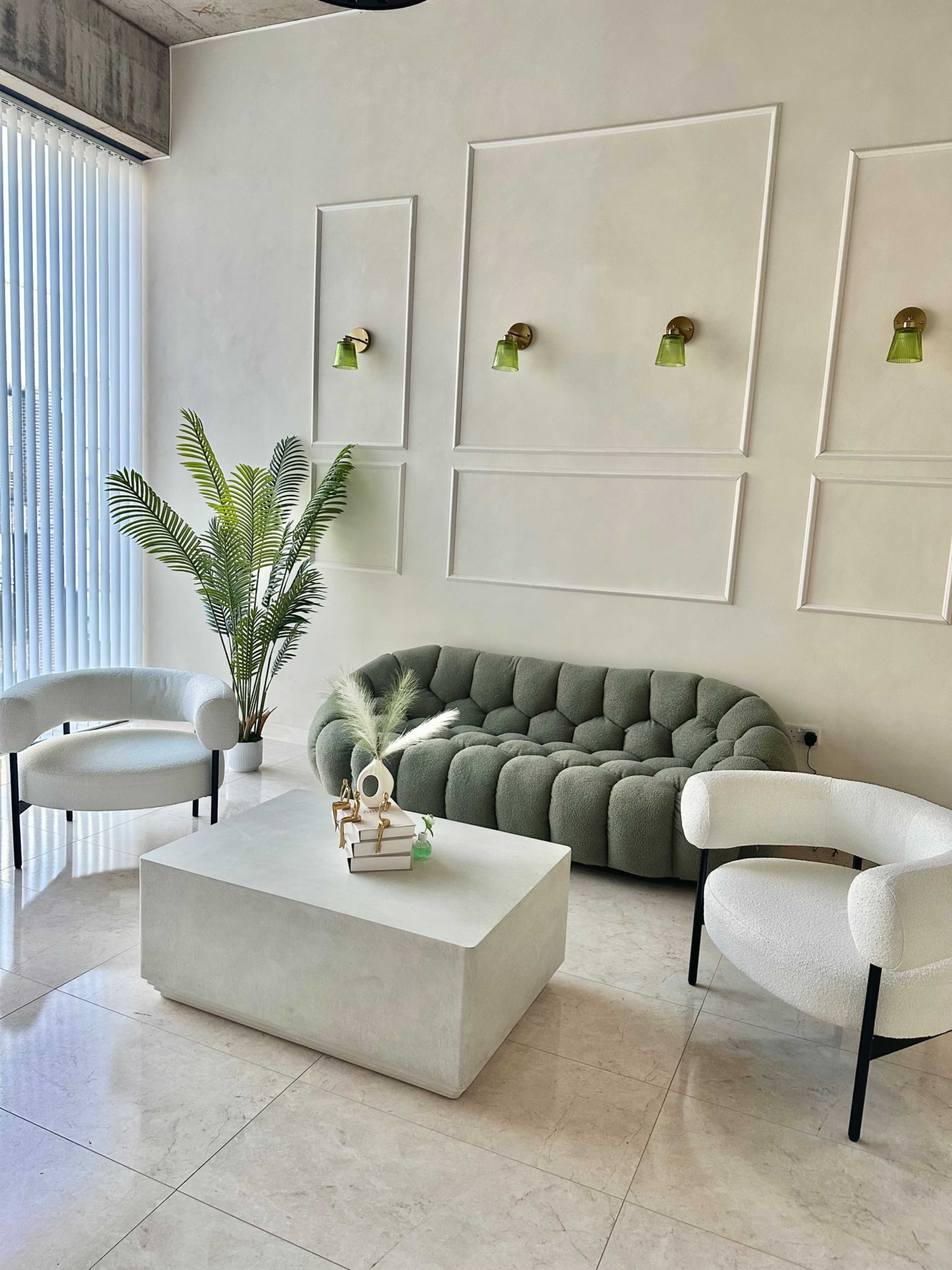The image shows a modern living area featuring a green tufted sofa, two white armchairs, a light-colored coffee table, and a potted plant against a textured wall with wall sconces.