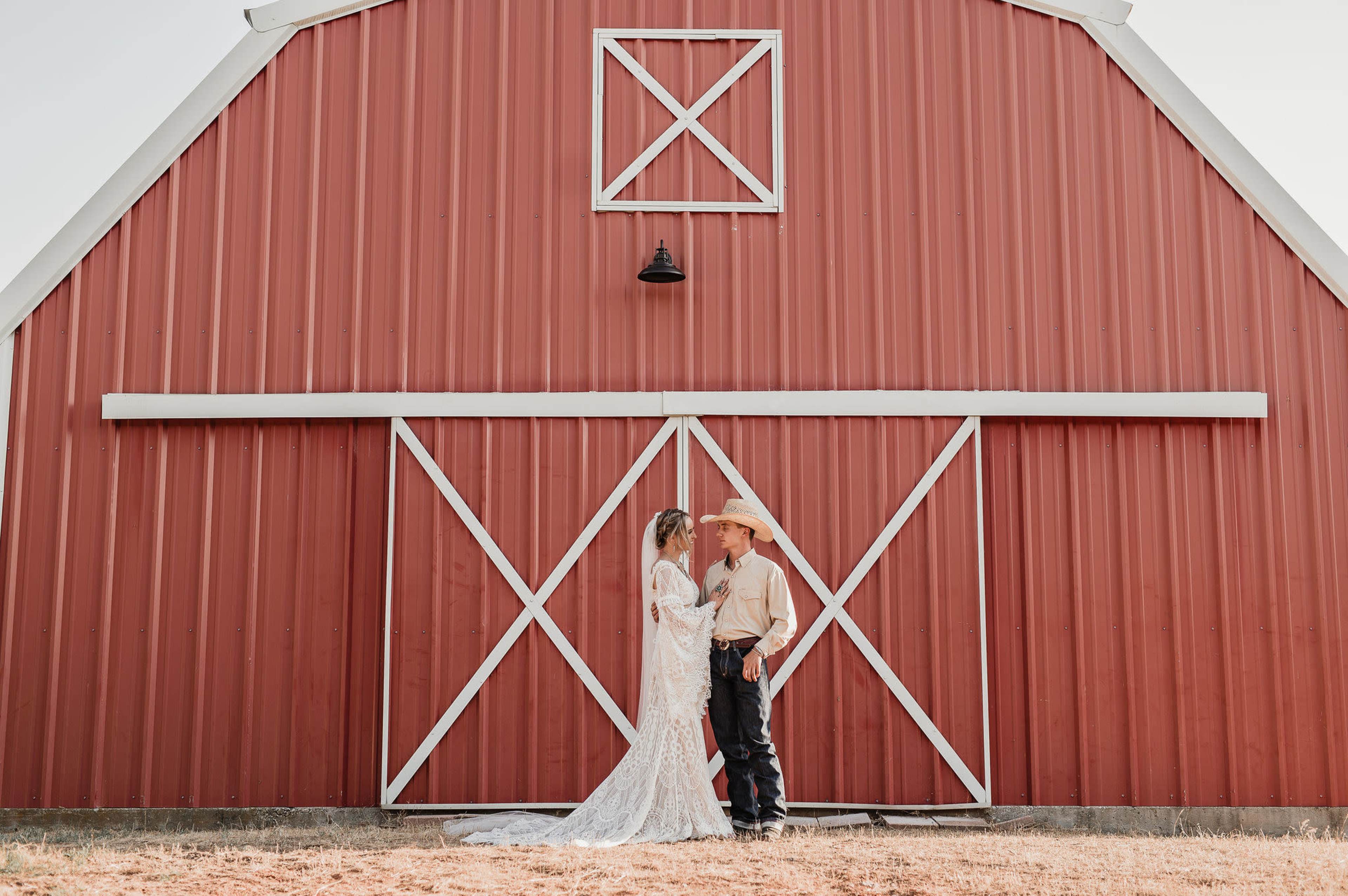 A couple stands closely together in front of a red barn, with the bride in a white dress and the groom in cowboy attire.