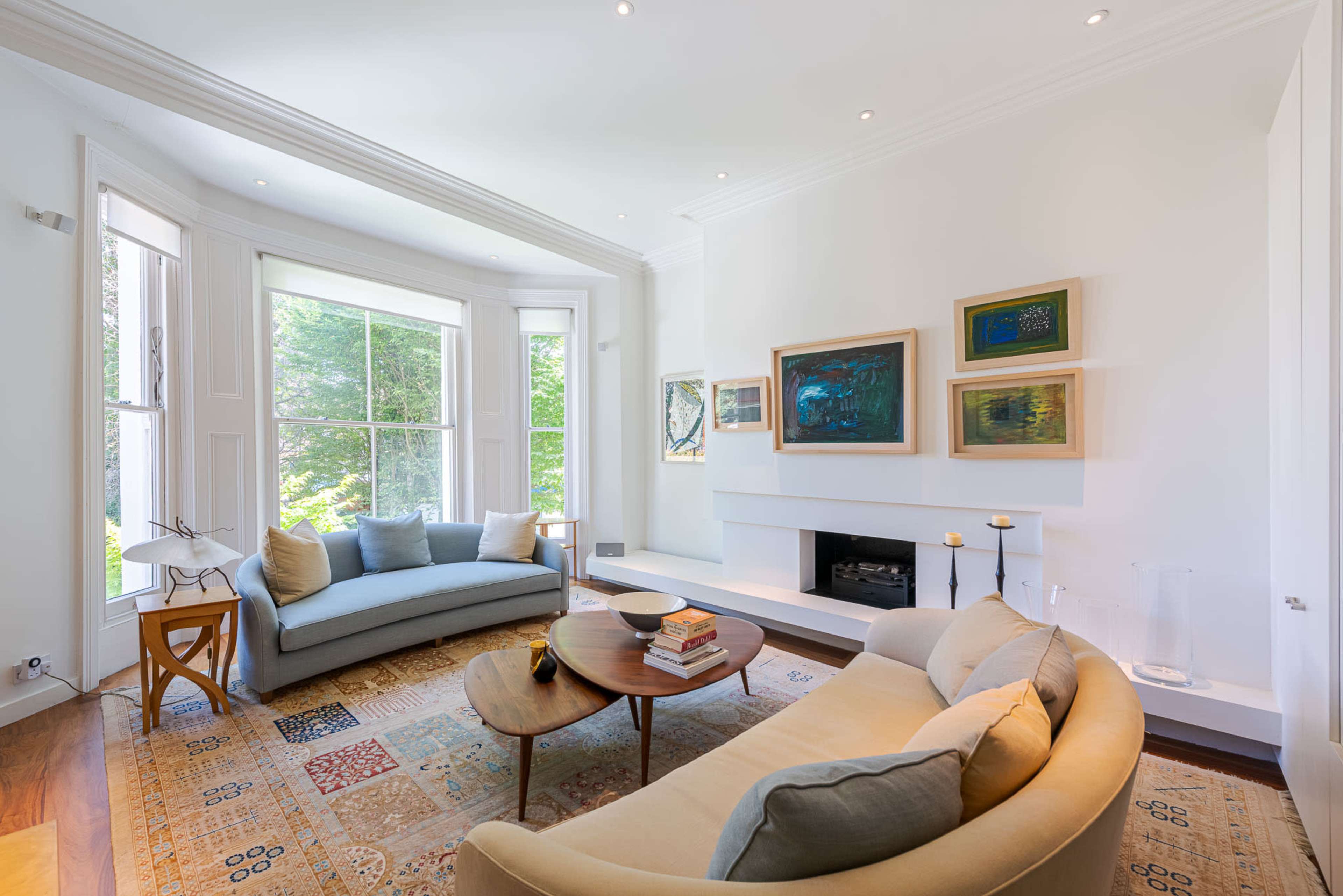A bright, spacious living room features two sofas, a round coffee table, and artwork displayed on the walls near a fireplace, with large windows allowing natural light to fill the space.