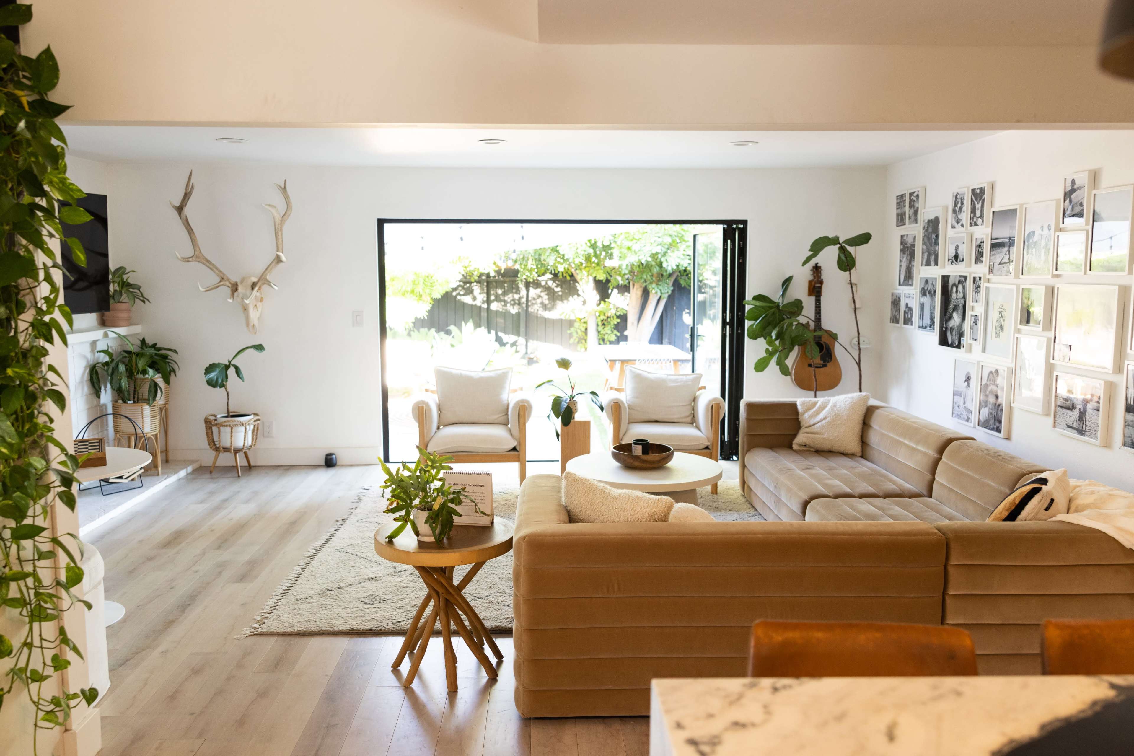A spacious living area features a beige sectional sofa, circular coffee table, and large glass doors that open to a backyard, adorned with plants and wall-mounted decor.