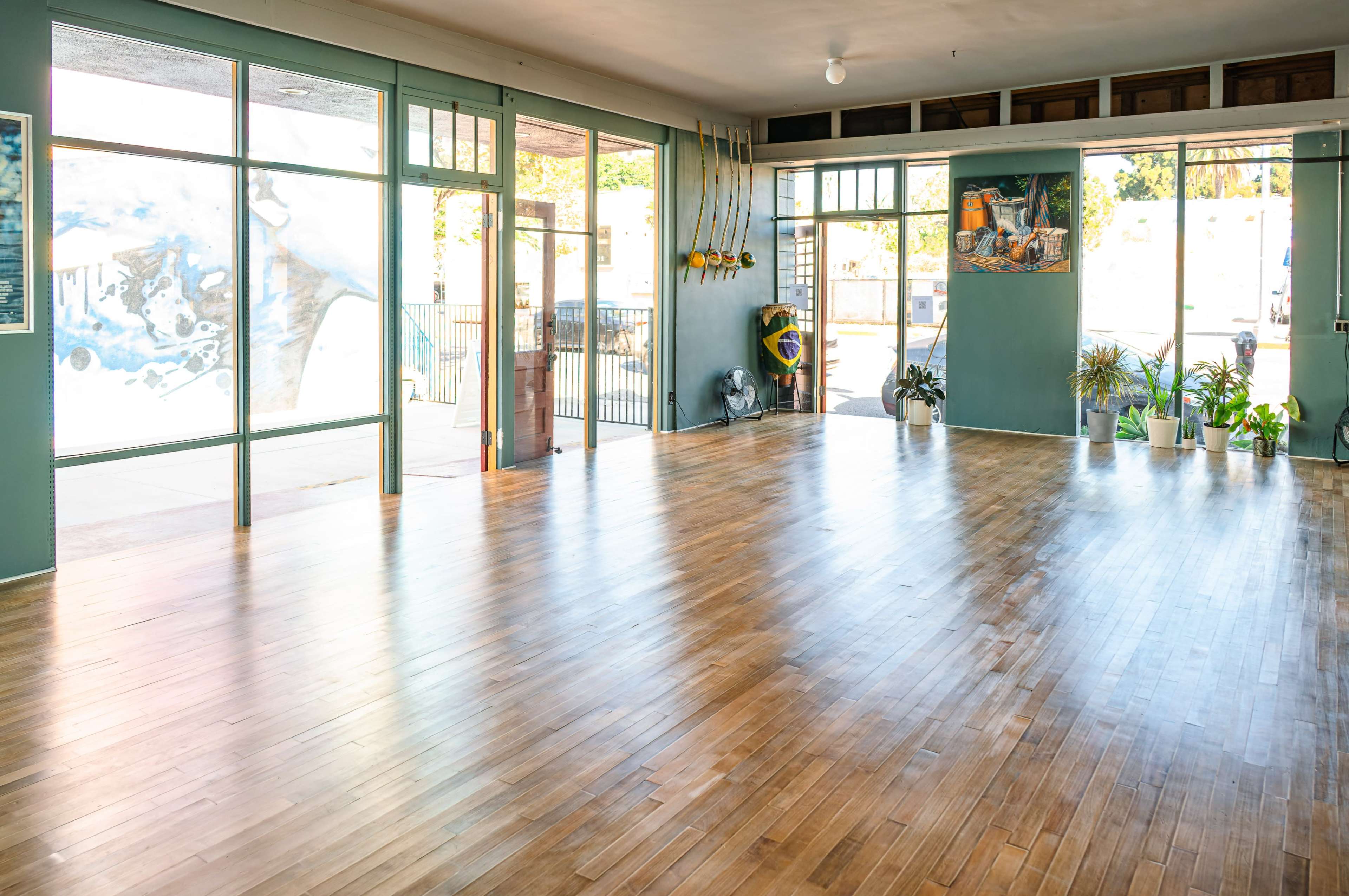 A spacious, empty room with wooden floors, large windows, and potted plants along the walls.