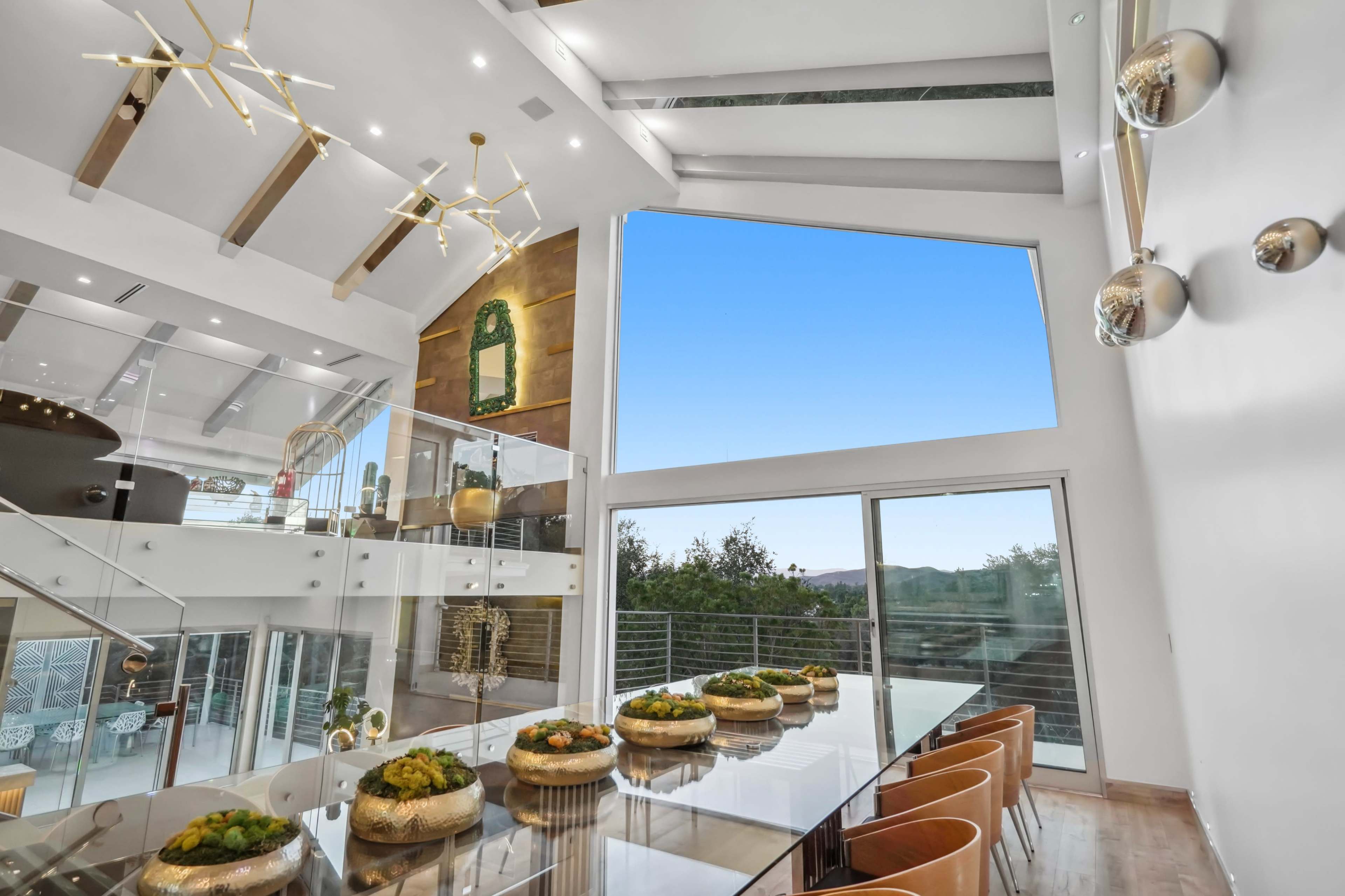 The image shows a modern dining area with a large glass table, stylish chairs, and an expansive window that provides a view of the sky and surrounding landscape.