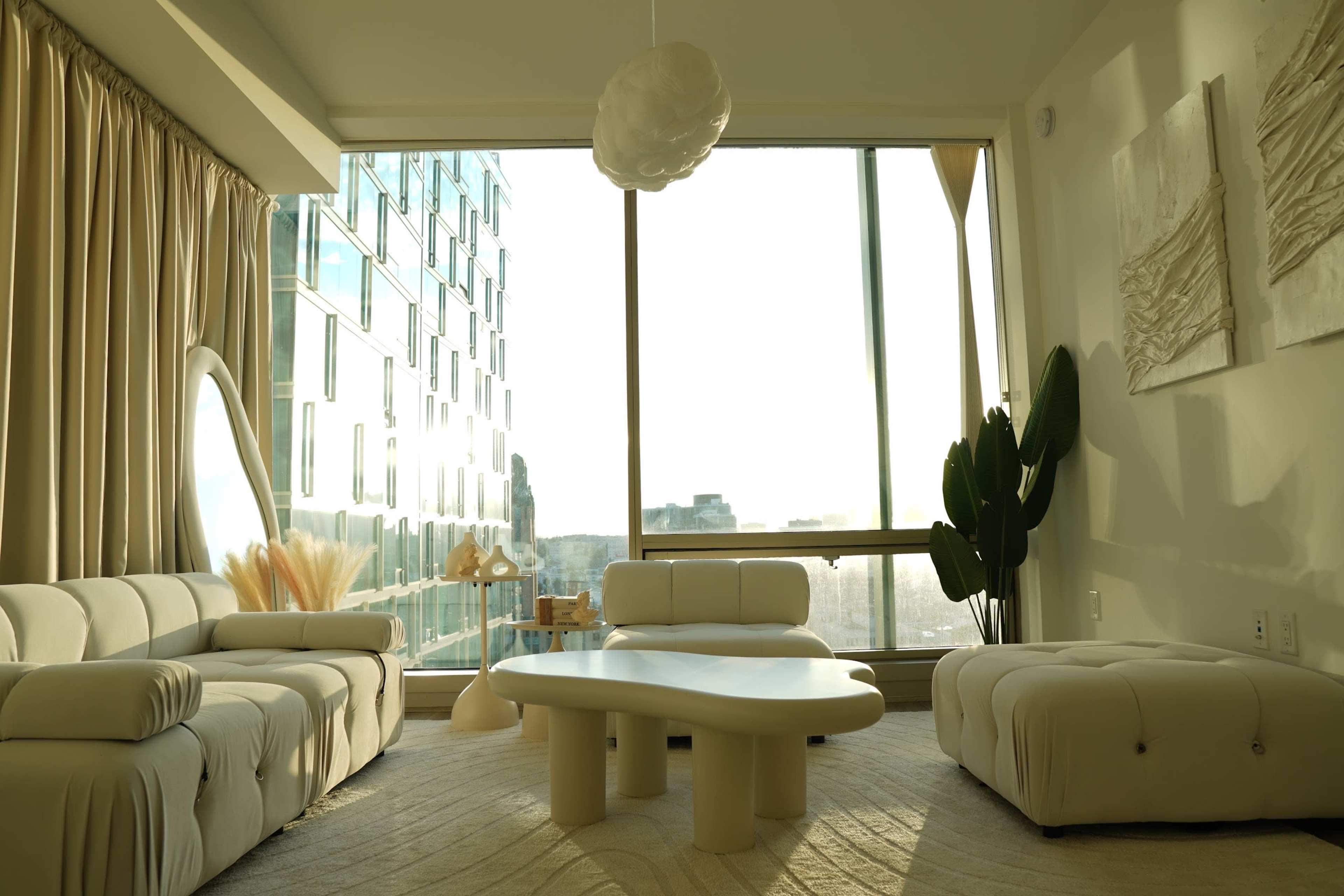 The image shows a modern living room with large windows, a minimalist coffee table, and beige upholstered furniture, set against a backdrop of a city skyline.