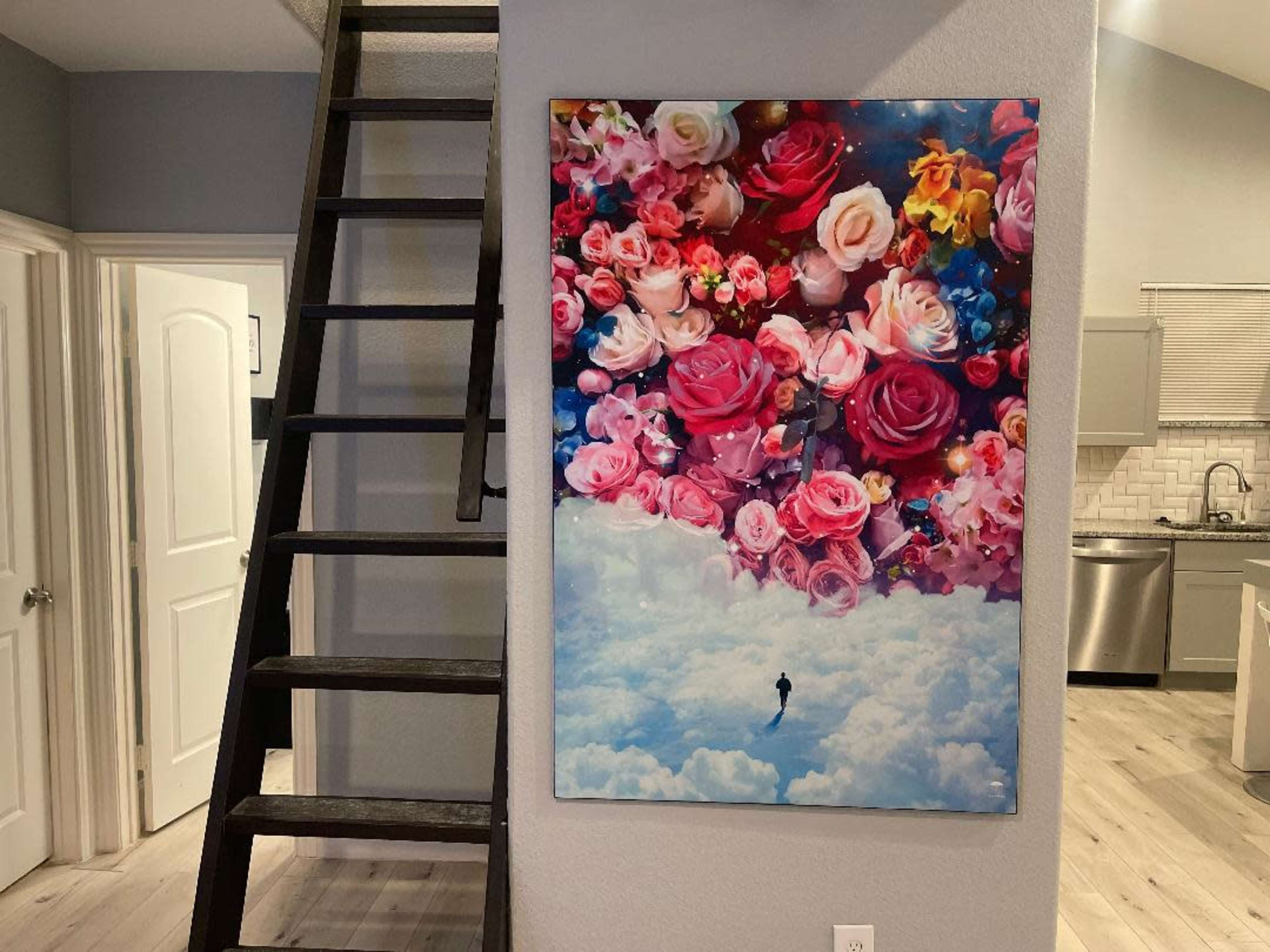 A large floral painting featuring roses and other flowers is mounted on a gray wall next to a wooden staircase in a well-lit interior space.