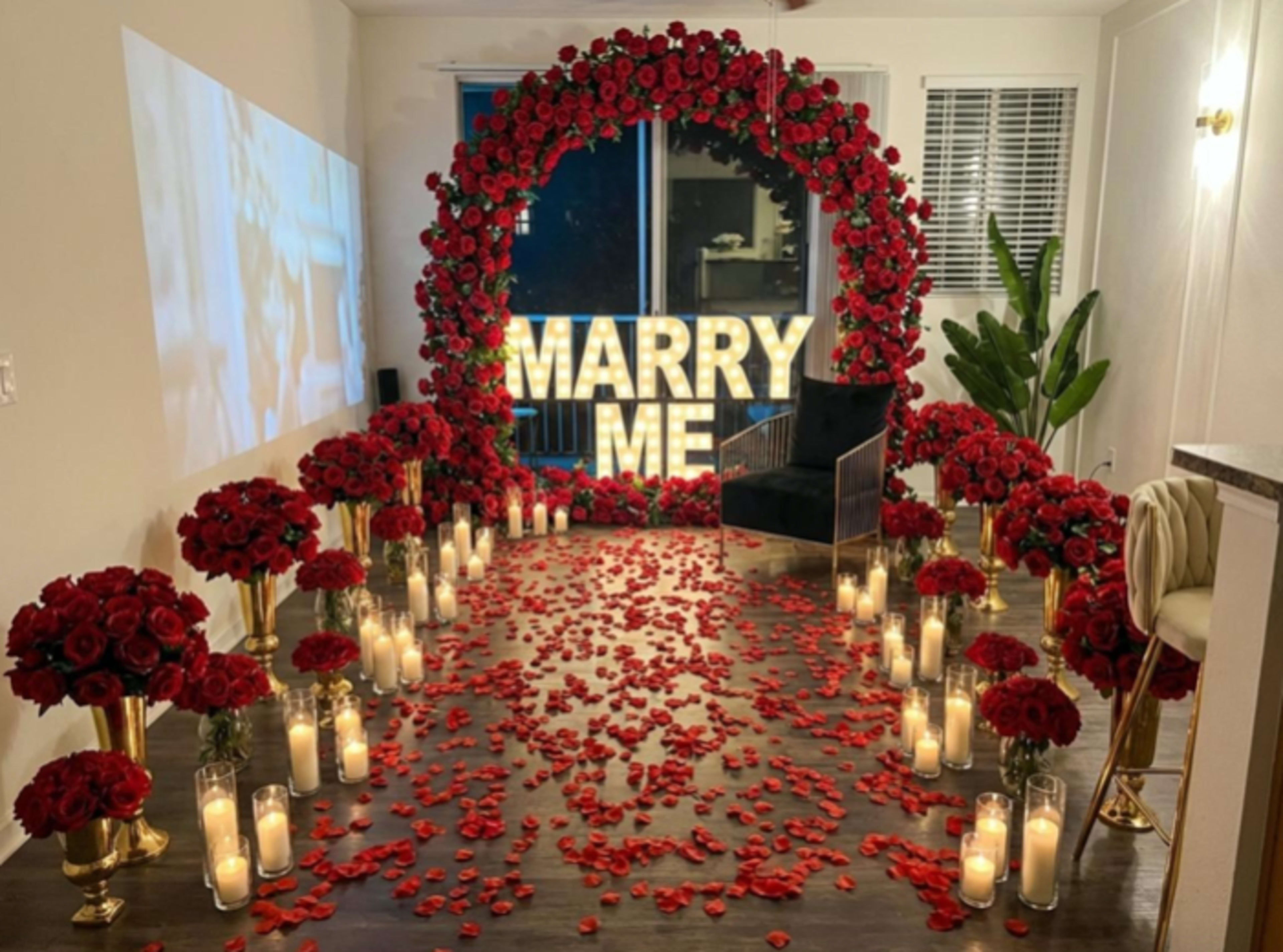The scene features a romantic proposal setup with large illuminated letters spelling "MARRY ME" surrounded by red rose petals, floral arrangements, and candles.