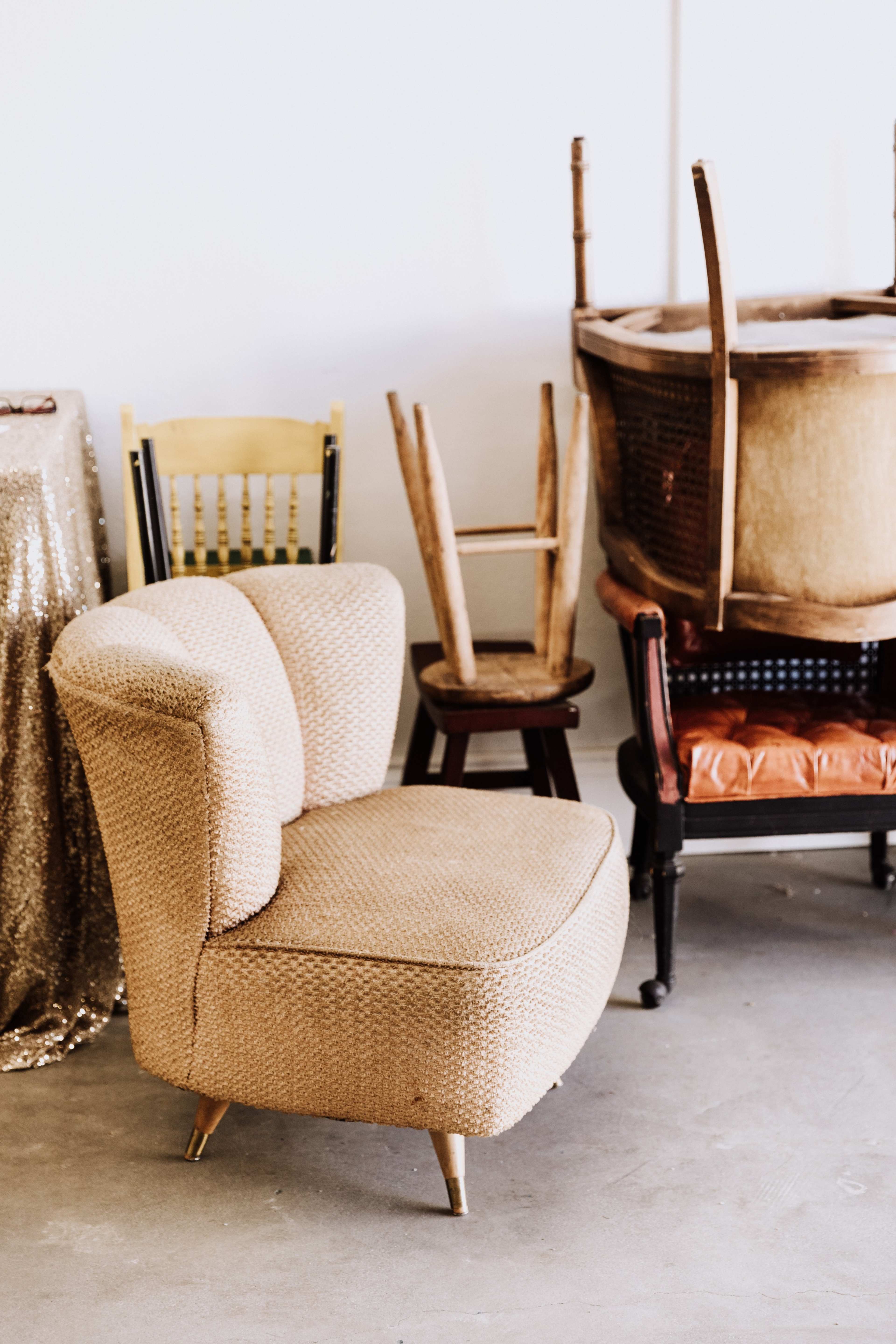 A textured beige chair sits next to various wooden chairs and a large wicker basket in a minimalistic indoor space.