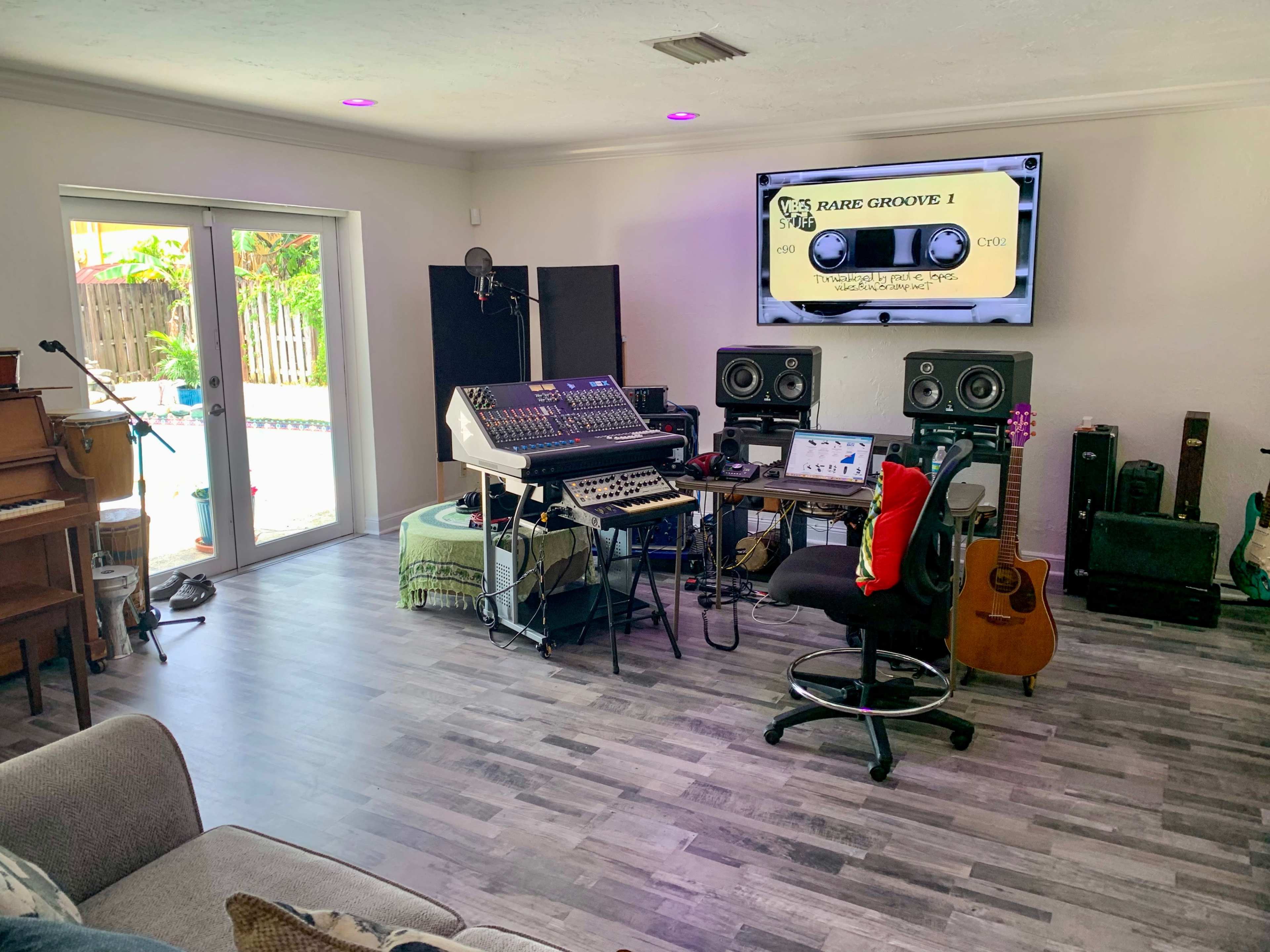 The image shows a home recording studio featuring an audio mixing console, musical instruments, and a large screen displaying digital audio software.