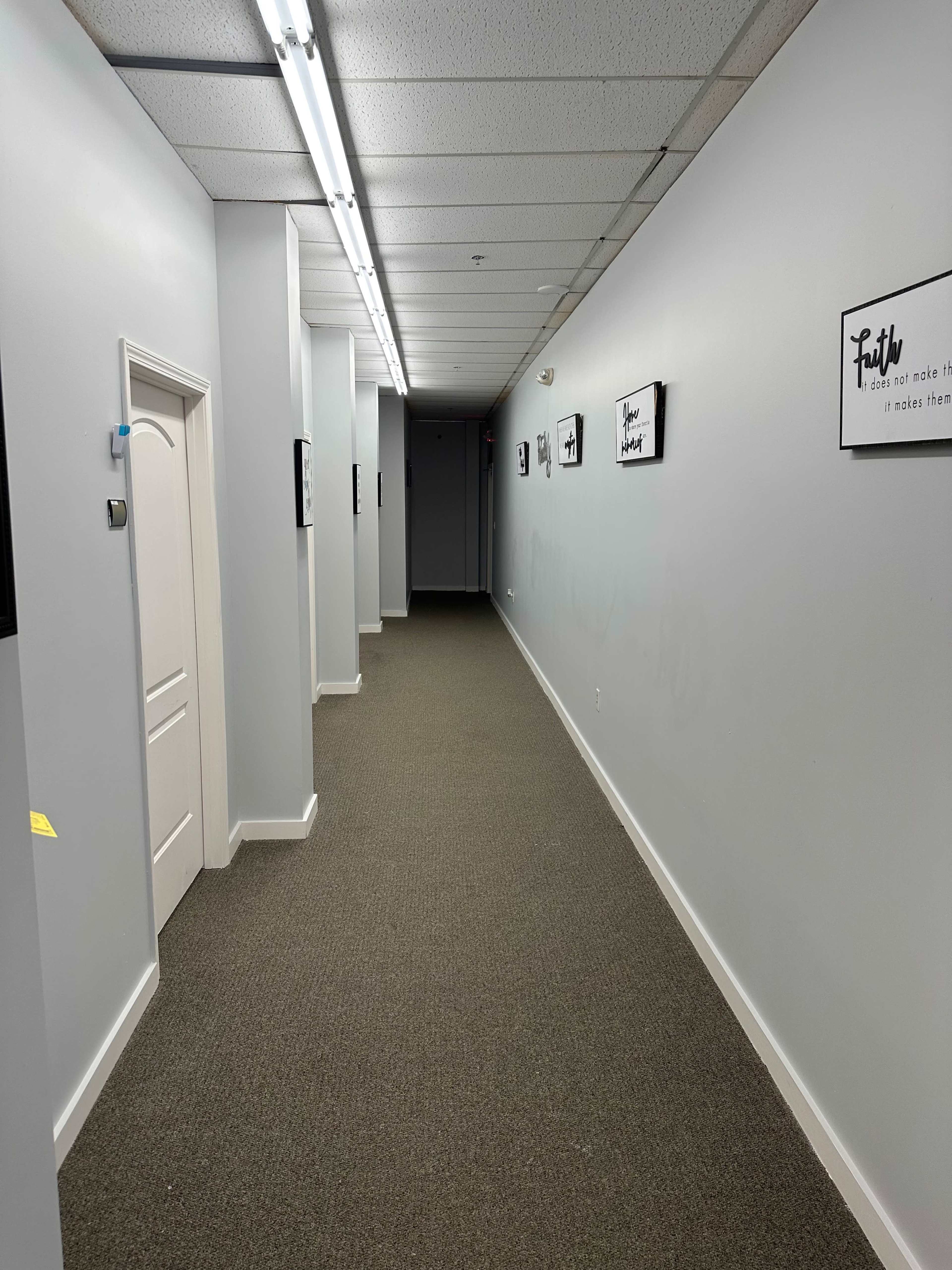 A long, narrow hallway with gray walls, a carpeted floor, and framed pictures hanging on either side.