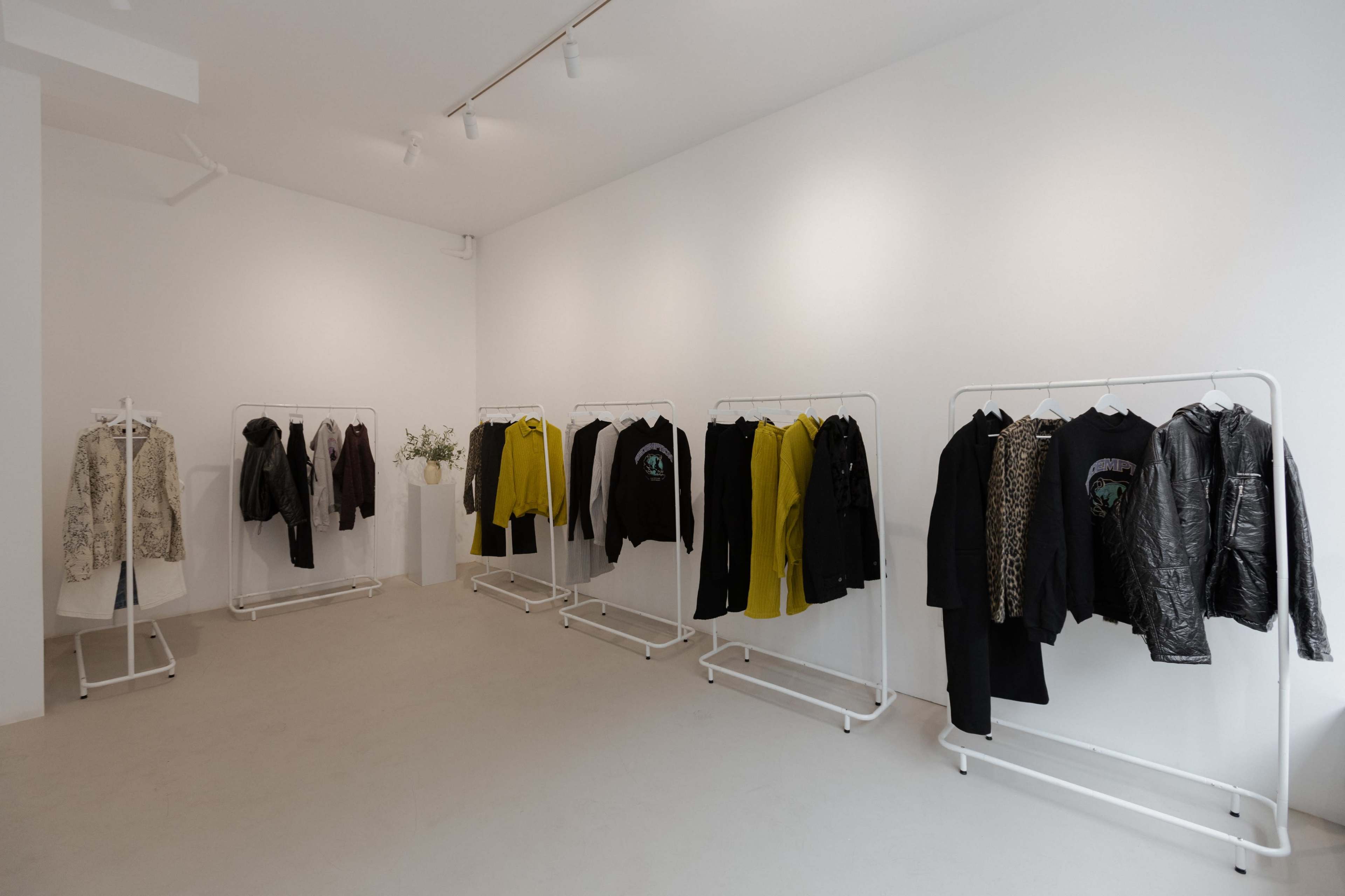The image shows a minimalist clothing store interior with racks displaying various jackets and tops in a range of colors, including black and yellow, against white walls.