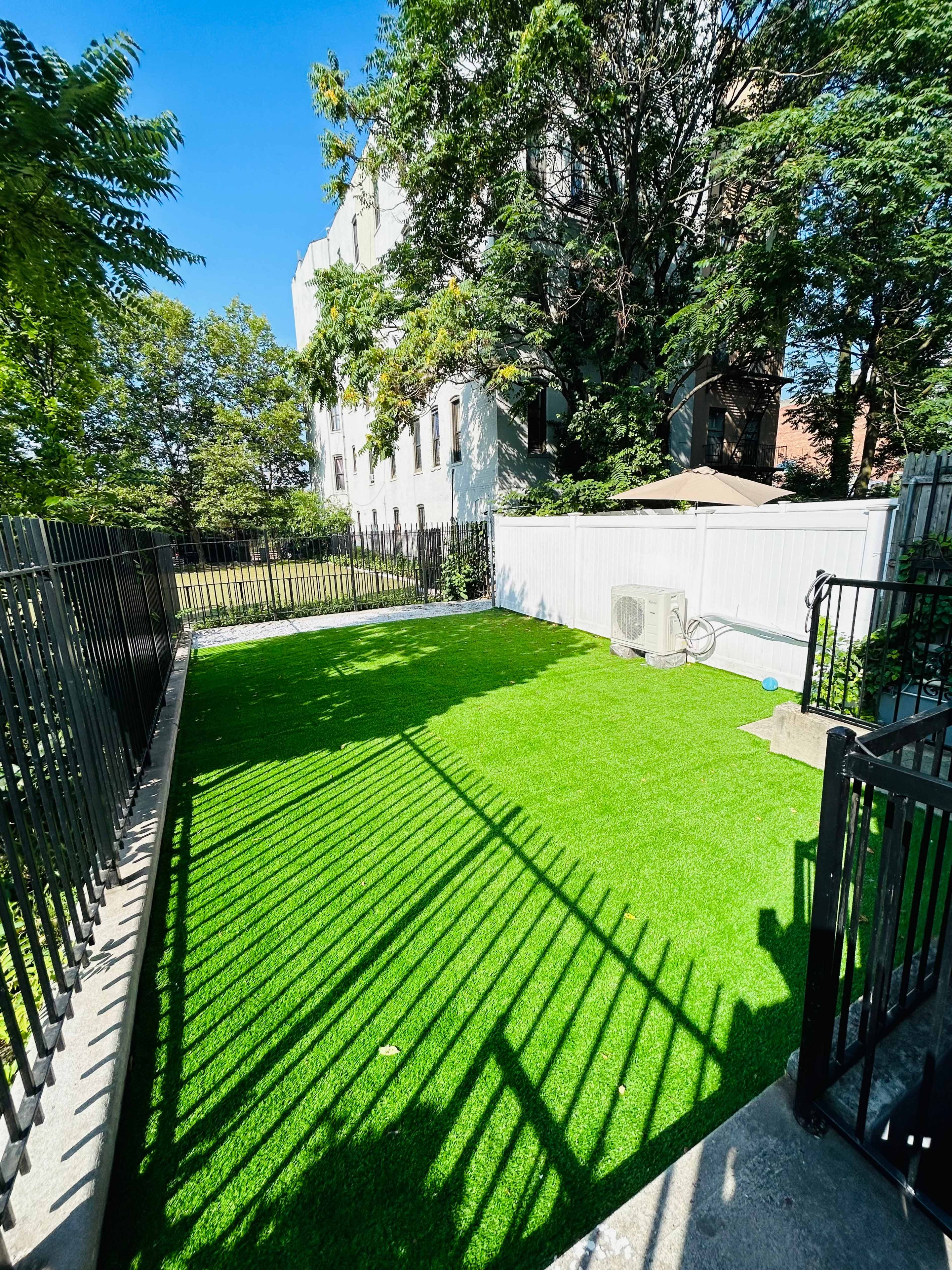 The image shows a small fenced yard with artificial grass, bordered by trees and a building in the background.