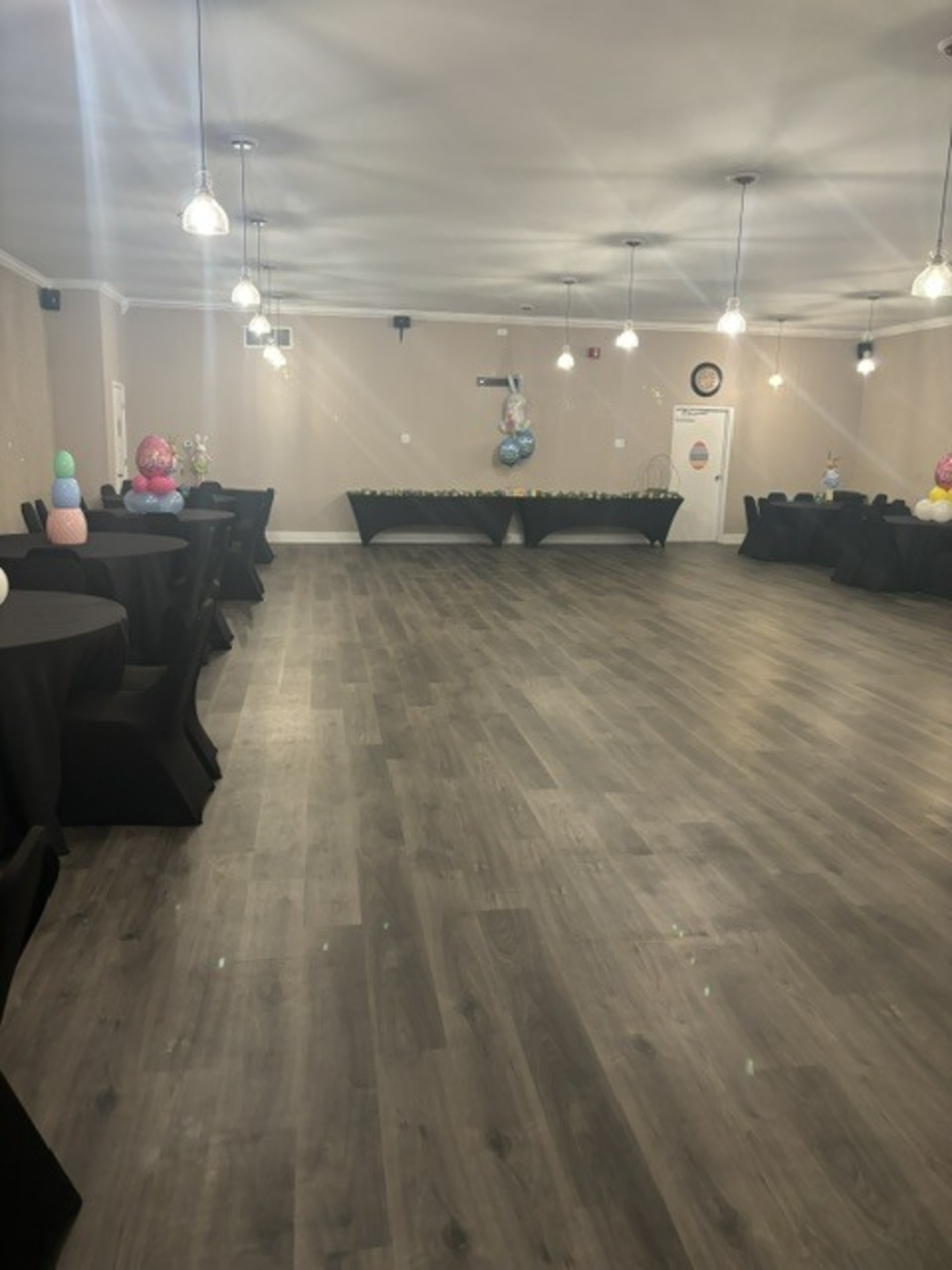 The image shows a spacious event hall with black tablecloths on round tables, decorated with balloons at one end.