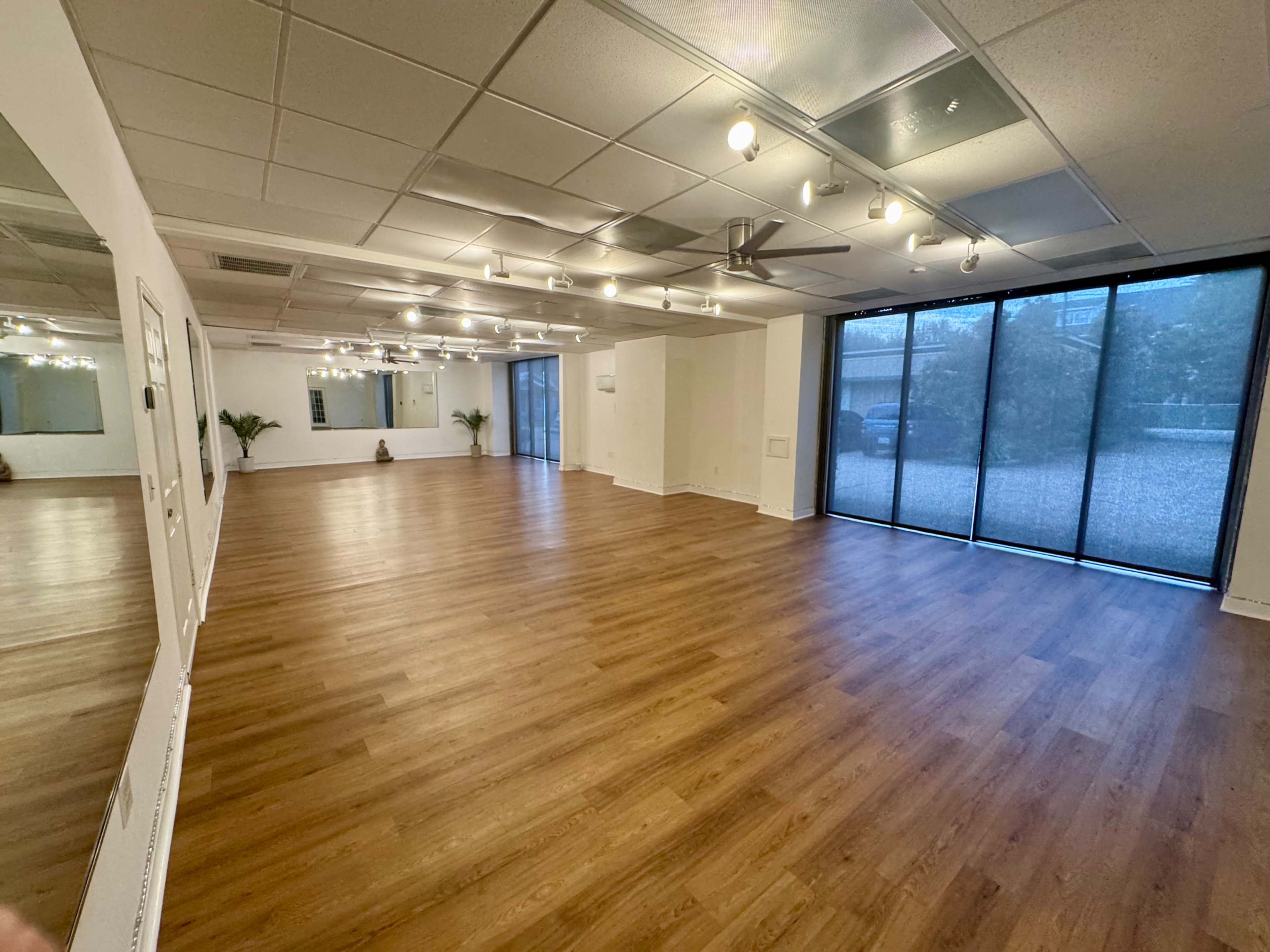 The image shows a spacious, well-lit room with wooden flooring, large windows, and minimalist decor.