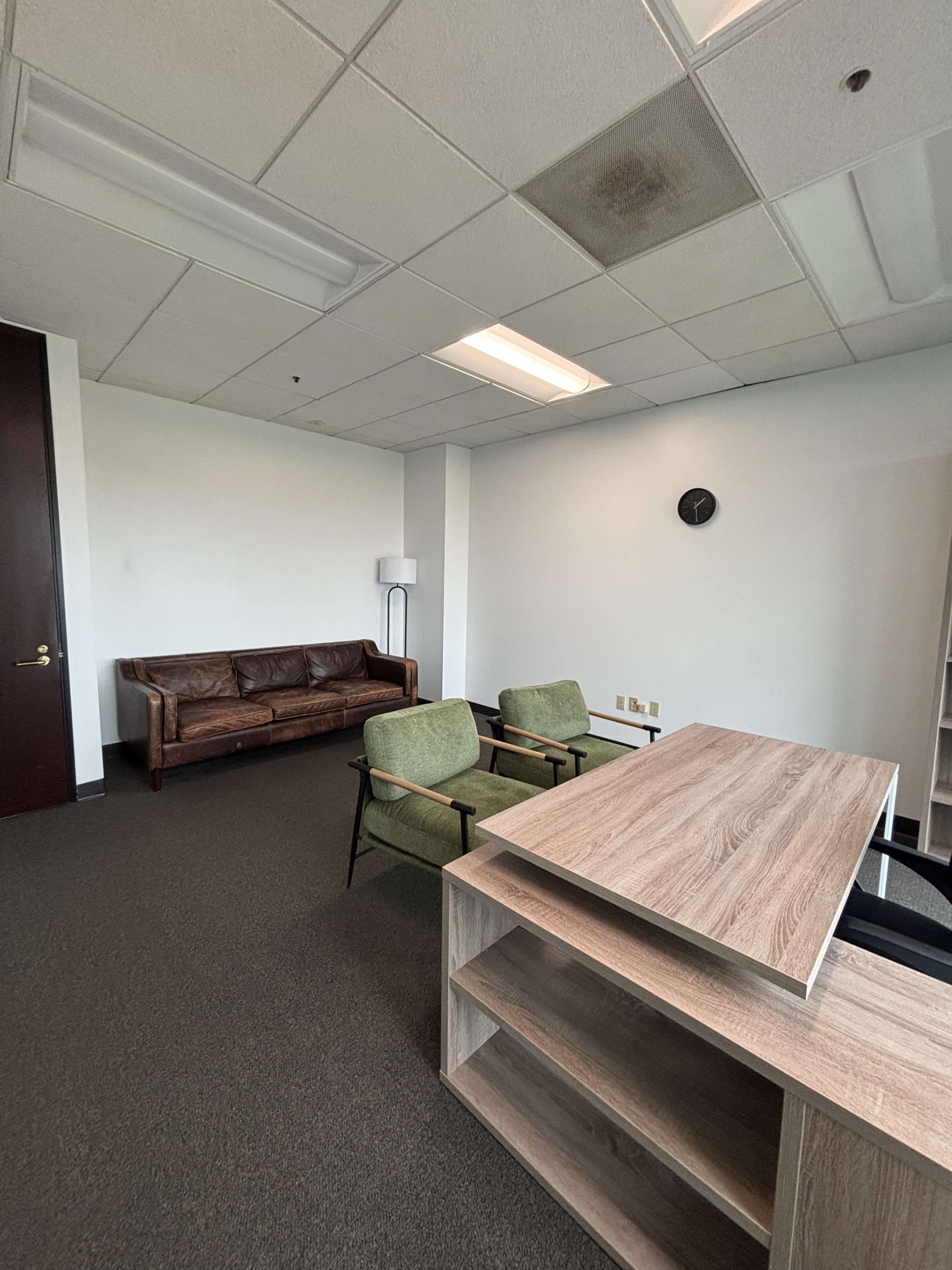 The image shows a spacious office with a brown leather sofa, two green chairs, a wooden desk, and a wall clock.