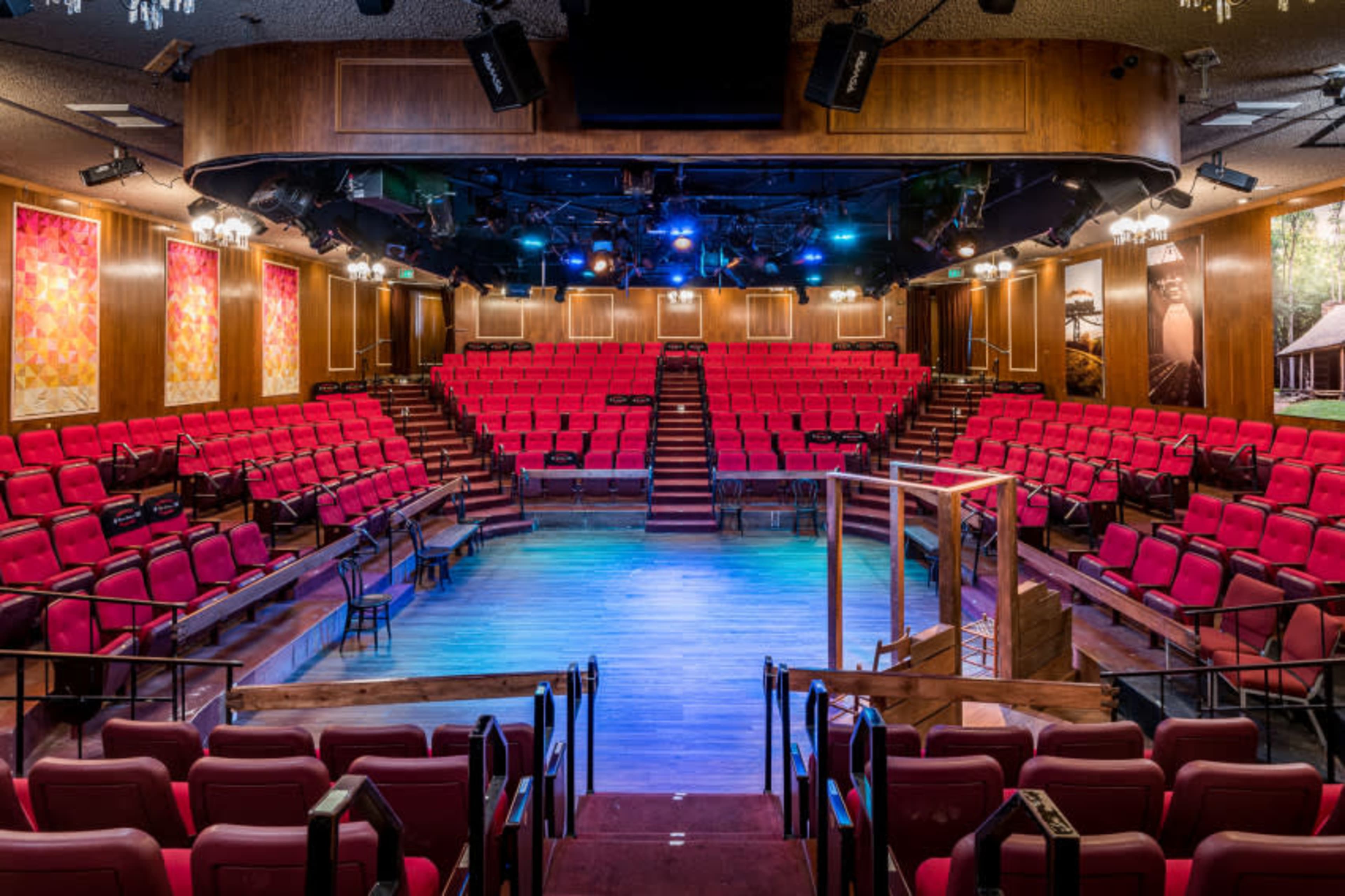 The image shows a theater with bright pink seating arranged in a semi-circular layout surrounding a stage, illuminated by colorful lights.