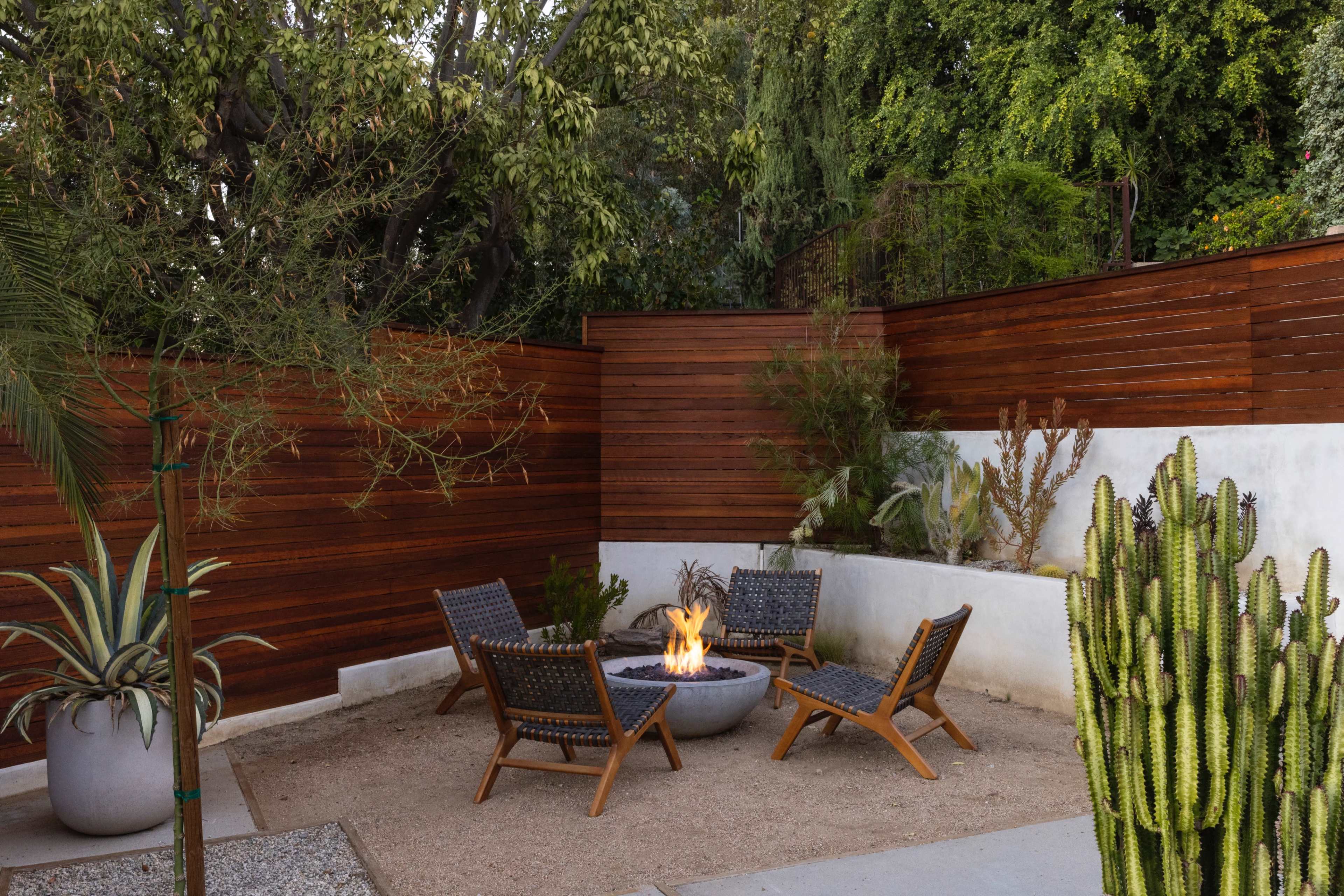A circular fire pit surrounded by four woven chairs is set in a gravel area, bordered by wooden walls and plants.