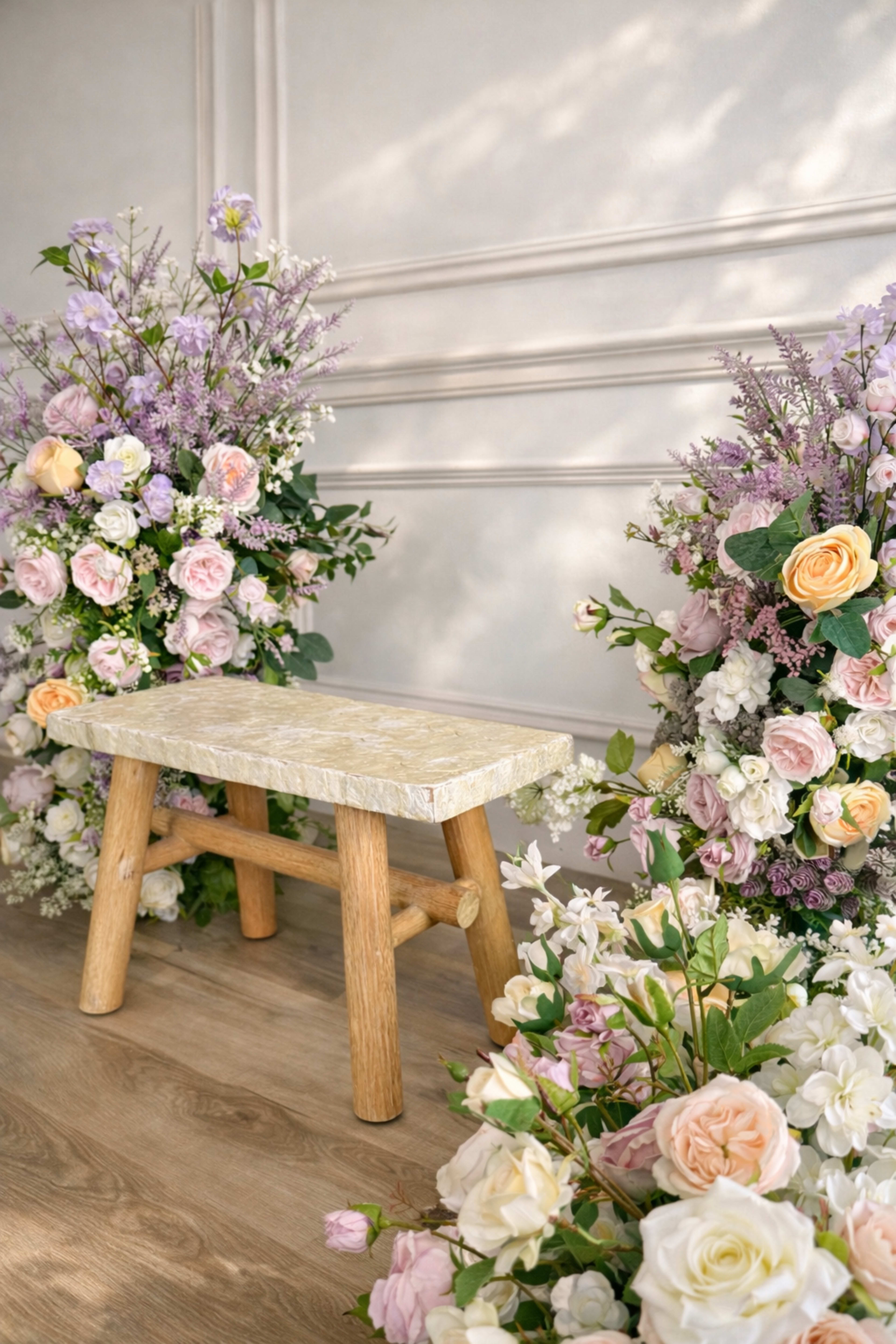A wooden stool is positioned between arrangements of pastel-colored flowers against a neutral wall.