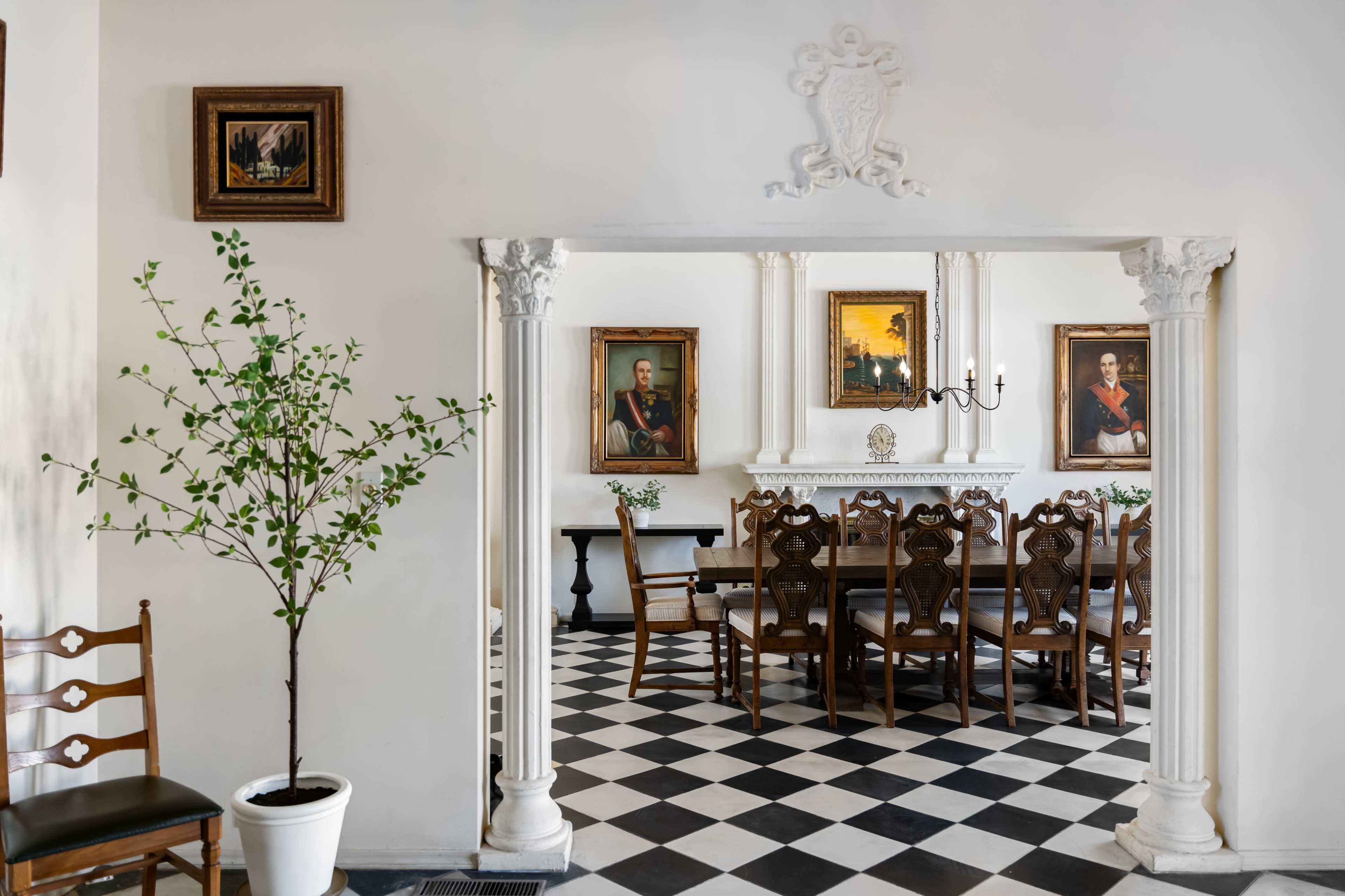 A spacious dining area features a long wooden table surrounded by chairs, with framed portraits on the walls and a potted plant in the corner.