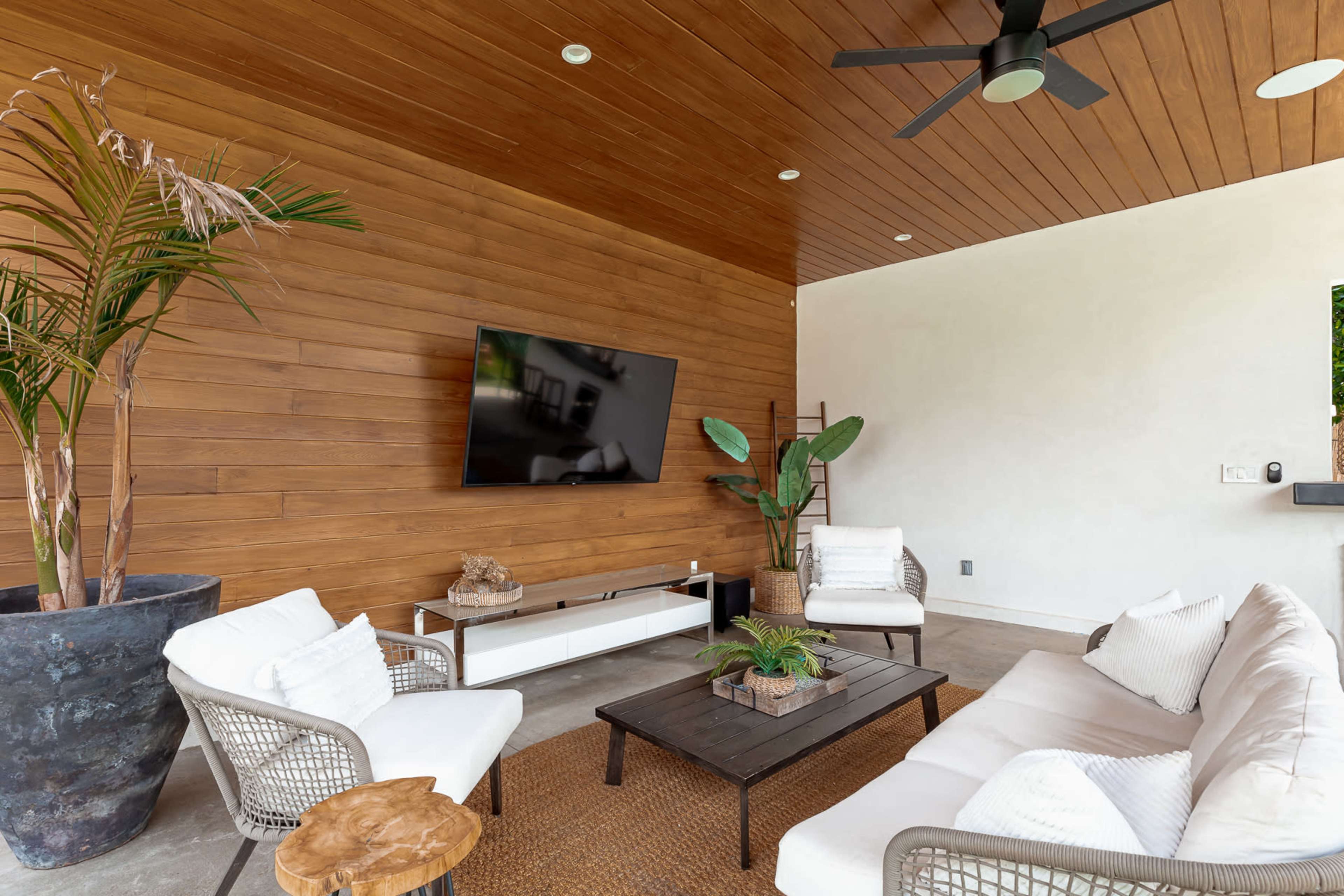 A modern outdoor living space featuring a wooden ceiling, a television mounted on the wall, and comfortable seating arranged around a coffee table.