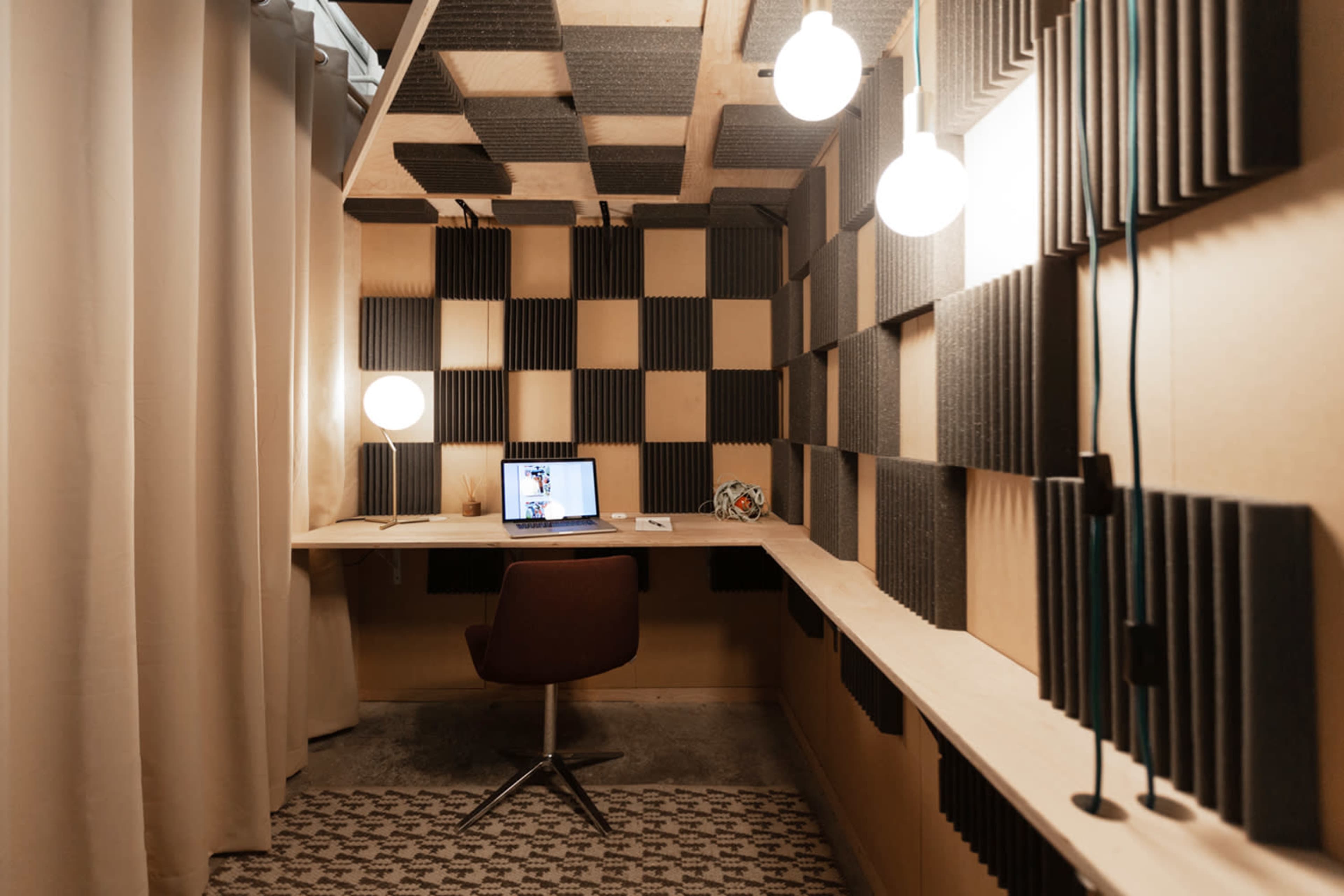 A small, soundproofed office space featuring a desk with a laptop, surrounded by patterned acoustic panels and illuminated by hanging light fixtures.