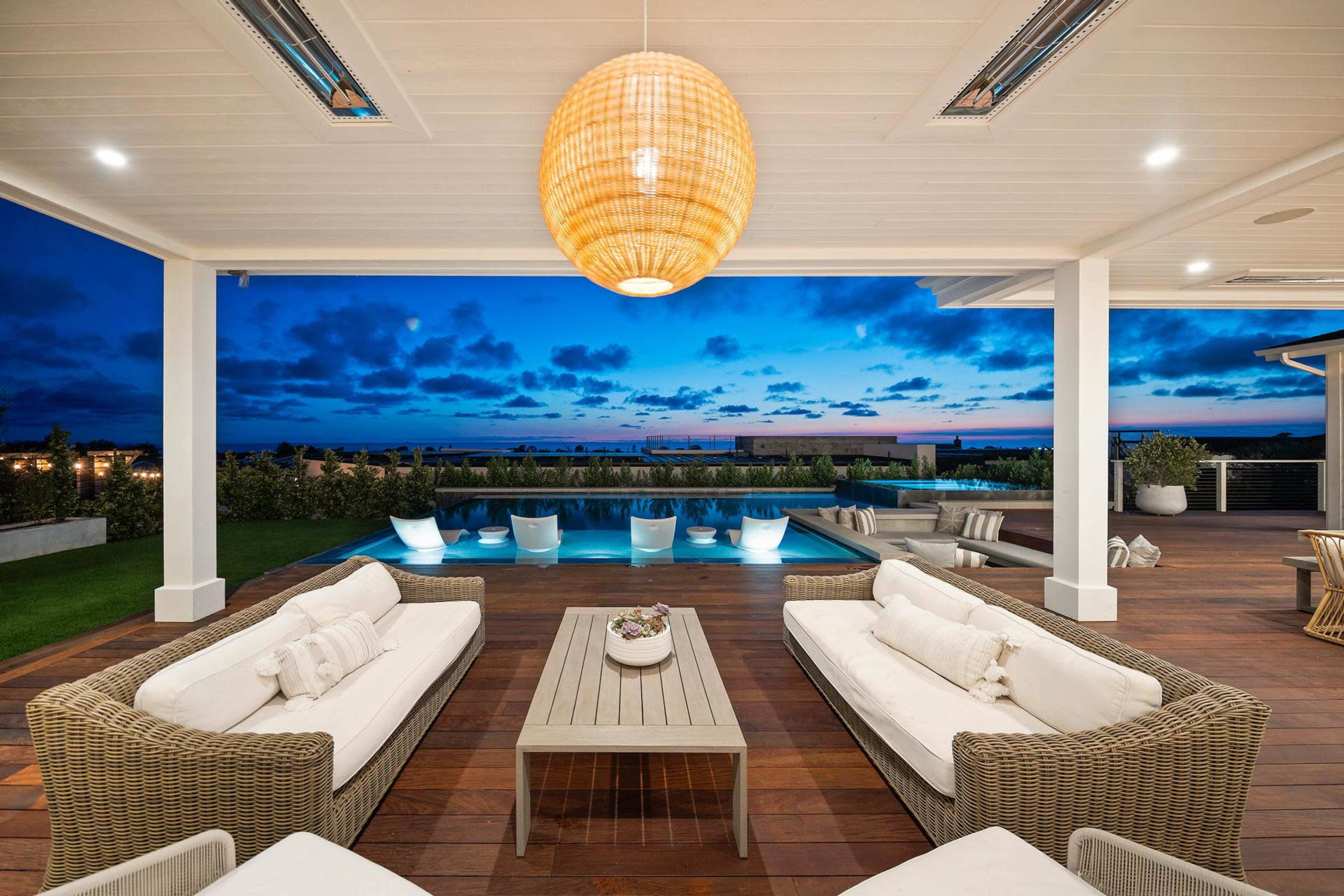 The image shows a modern outdoor living space with wicker furniture arranged around a wooden coffee table, overlooking a pool under a twilight sky.